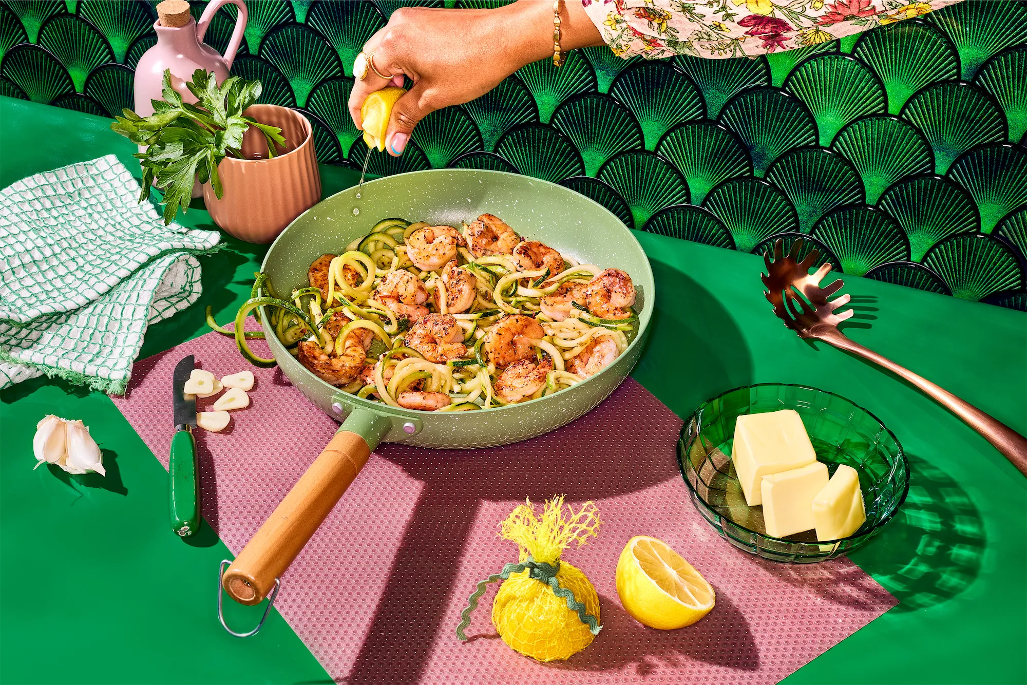 Pescanova brand shrimp and zucchini noodles in a ceramic pan on vibrant green backround and pink placemat with fresh lemon parsley and cubes of butter