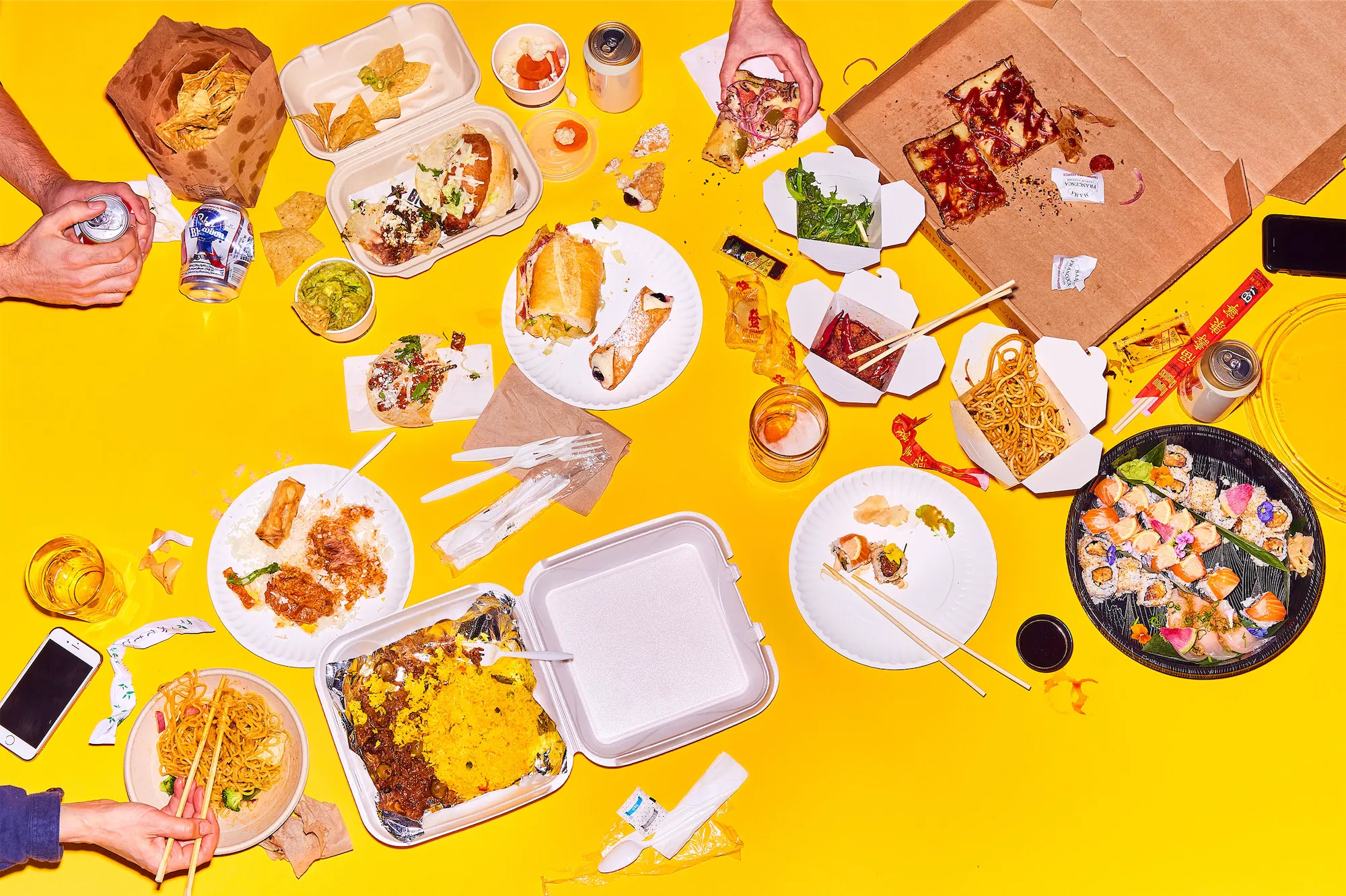 a messy chaotic scene of different take out foods and hands interacting on a yellow surface