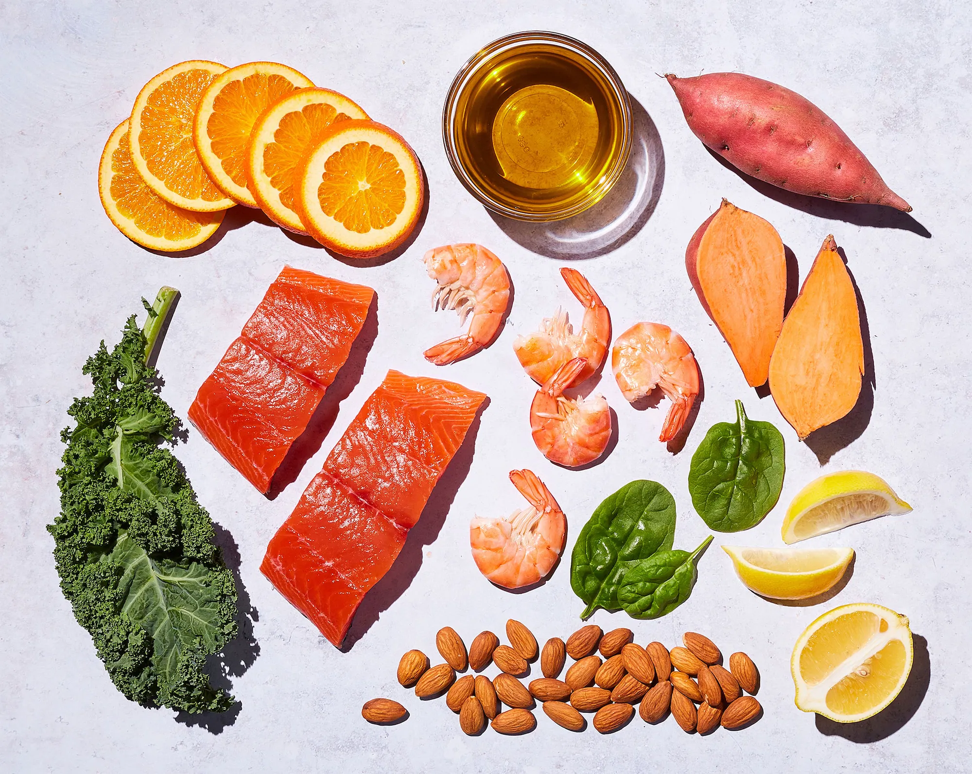 an arrangement of healthy ingredients salmon kale shrimp sweet potato almonds lemon spinach orangeson a lilac surface