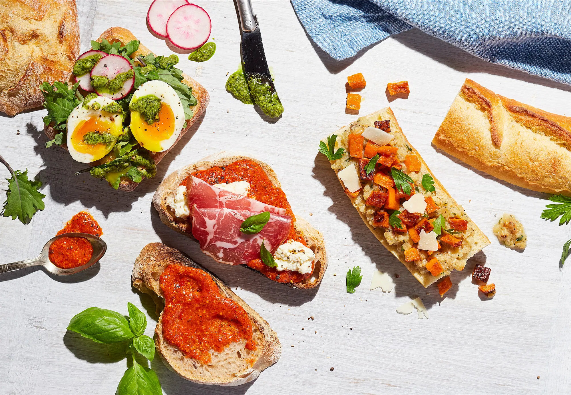 armanino brand foods overhead scene of sandwiches in process with charcuterie red pesto soft boiled eggs fresh basil french baguette on white surface blue napkin