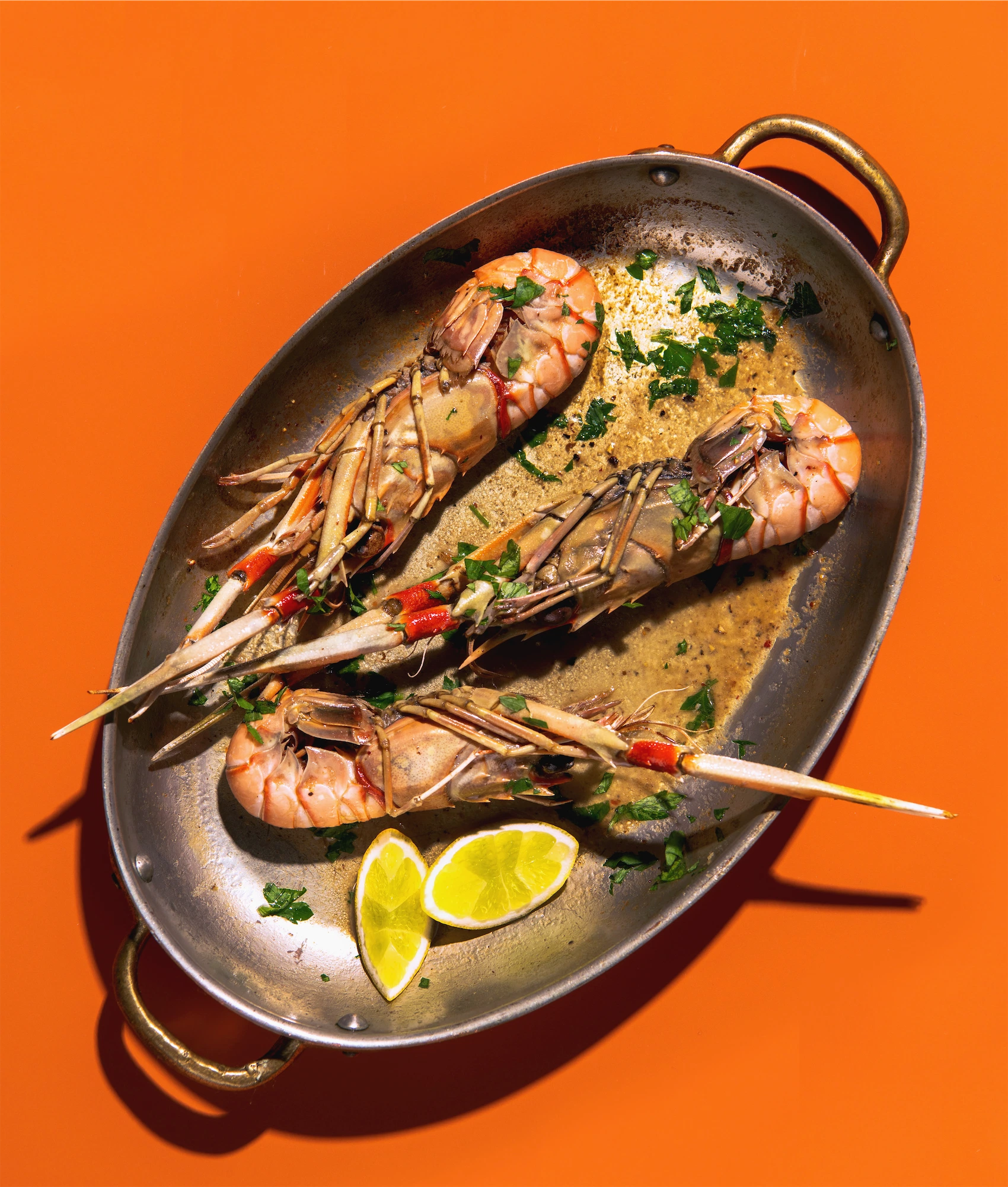 three langoustines in a french pan on an orange background with lemon wedges