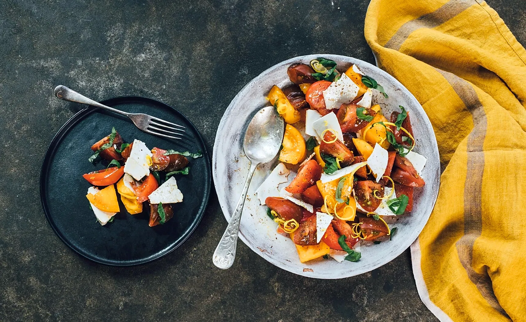 horizontal image of ricotta salata and tomato salad in a white bowl and on a black plate with a yellow napkin on dark grey surface