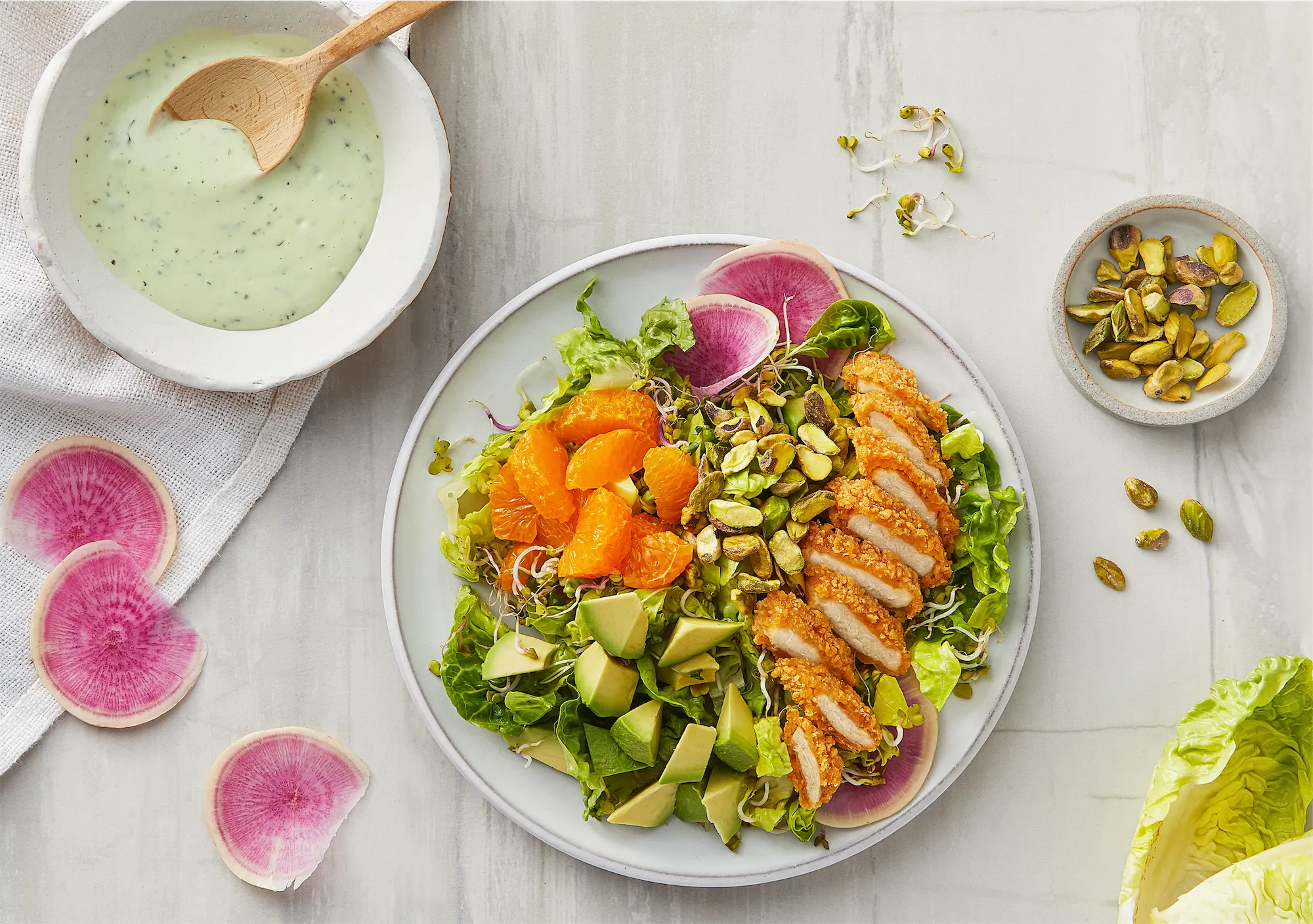 breaded chicken sliced on a white plate with mixed salad of lettuce avocado oranges pistachios and radish a creamy herb sauce sits upper left