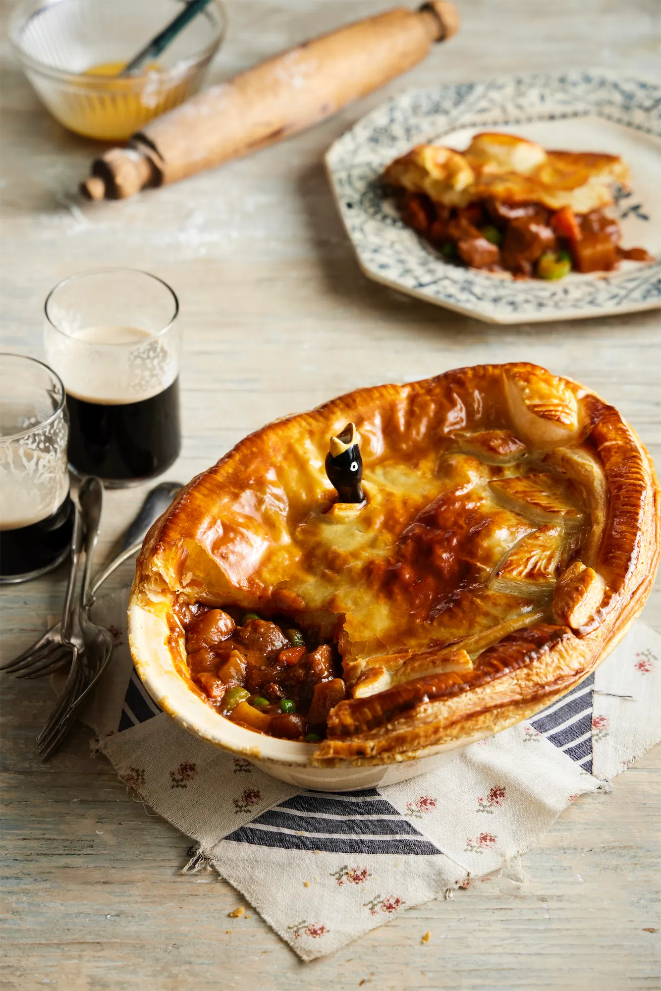 oval pie dish containing a beef and guiness pie with puff pastry and a pie bird a rolling pin and a patchwork placemat