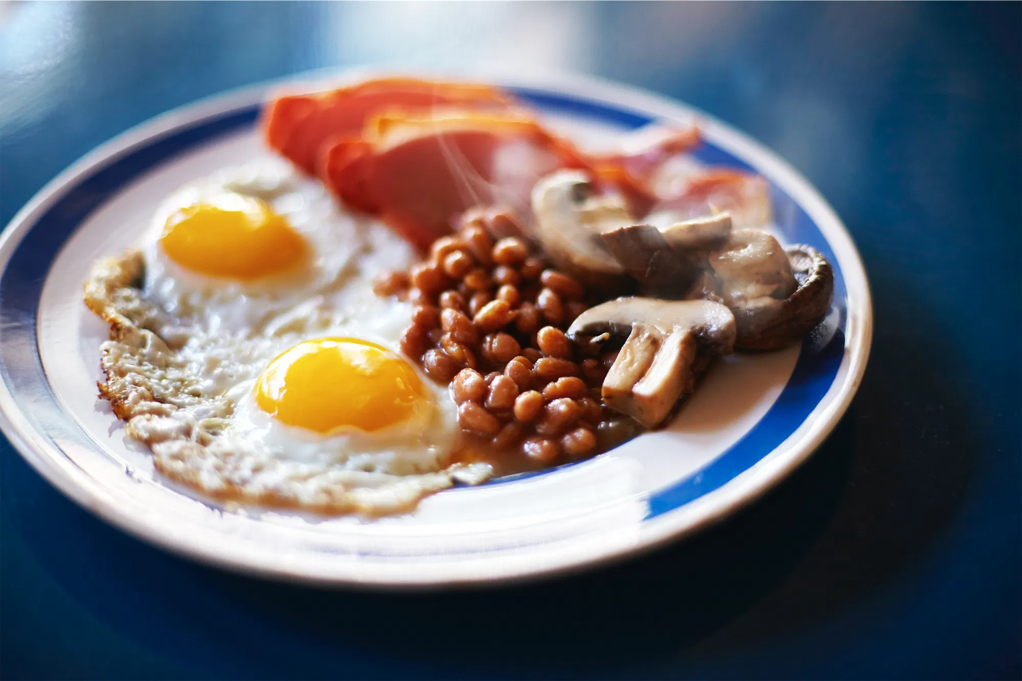 a blue and white striped plate of english style breakfast with two fried eggs baked beans bacon and mushrooms on a blue surface