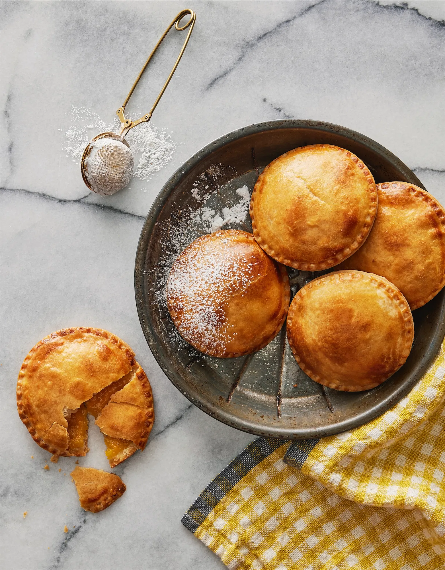 pie pan containing 4 round pastry pies and another pie on the marble surface broken into plus a sugar sifter upper left and a yellow check napkin lower right