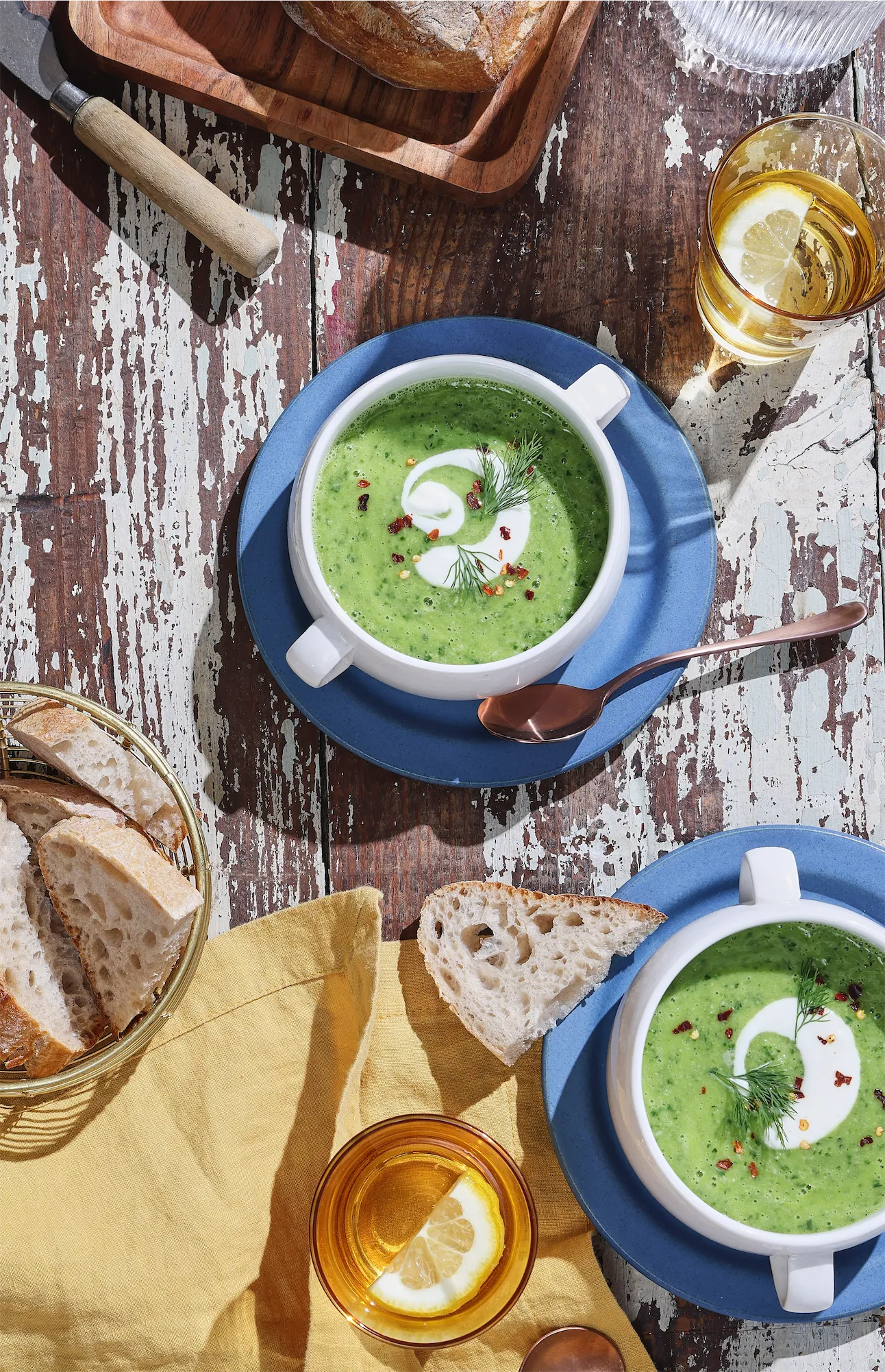 a sunny scene with two bowls of green soup and swirls of cream on a rustic table with yellow napkin and slices of bread