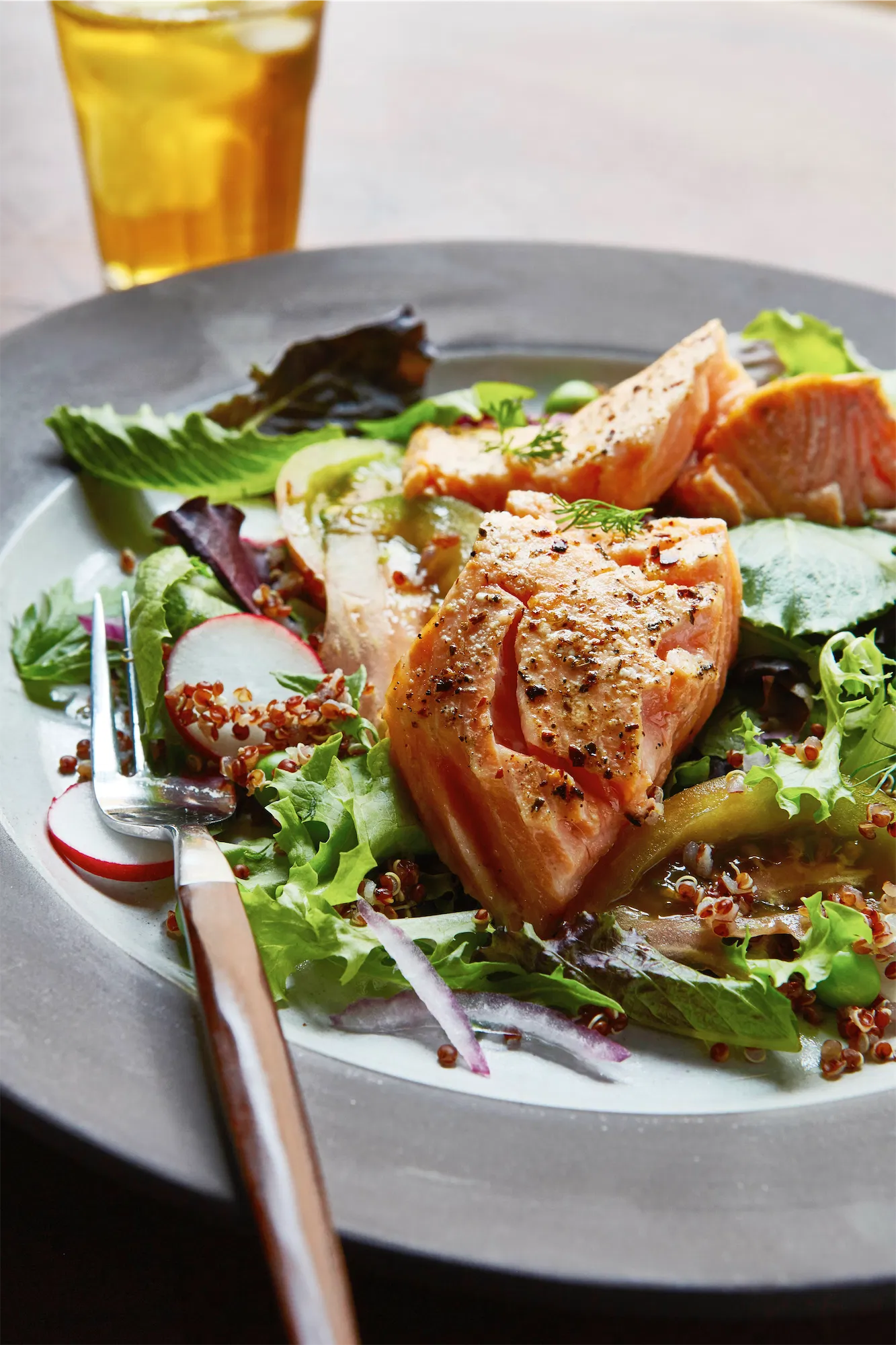 a white polate with flaky salmon on a bed of lettuce a wood handled fork and a yellow glass in the backgroound