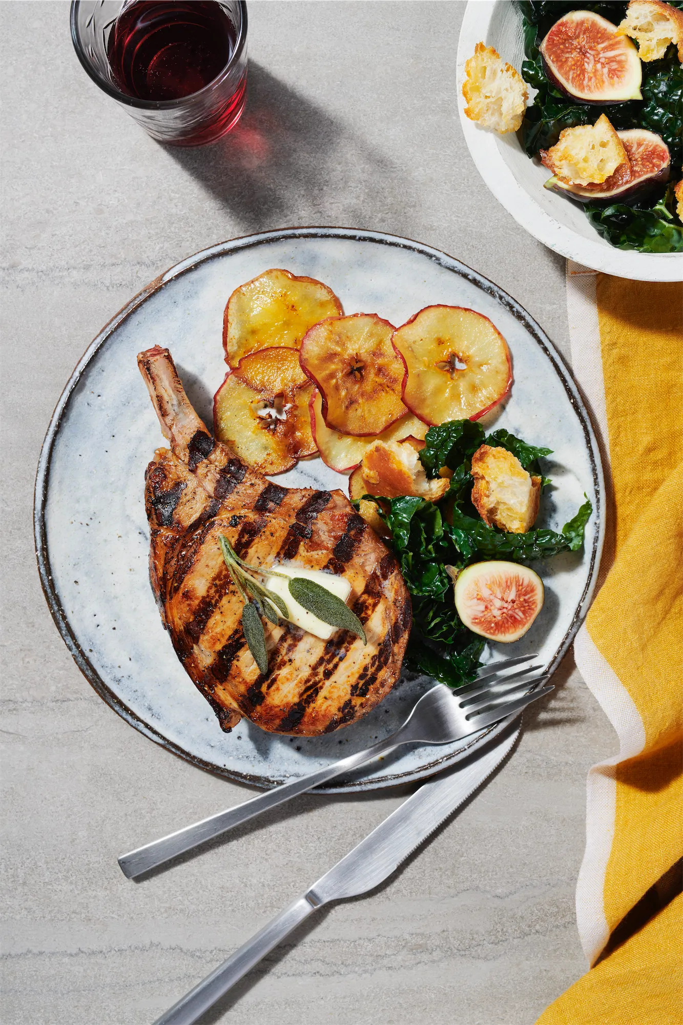 an overhead of a whit eplate with grilled pork chop garnished with sage and sliced apples a glass of red wine and a yellow napkin to the right