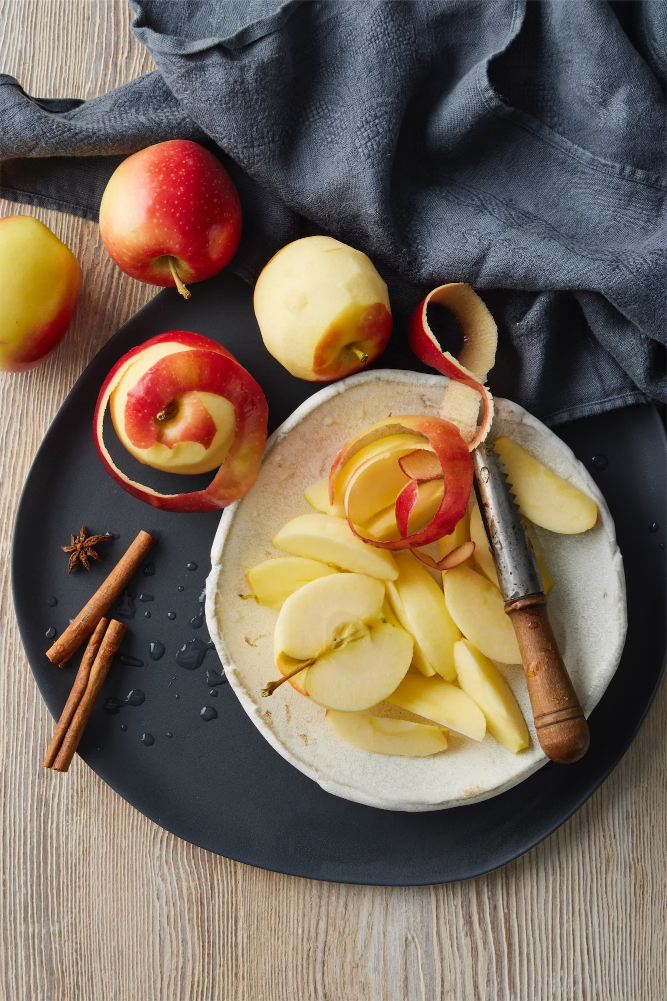 an overhead of a white plate on a dark grey platter with apples being peeld and cored and sliced for apple pie