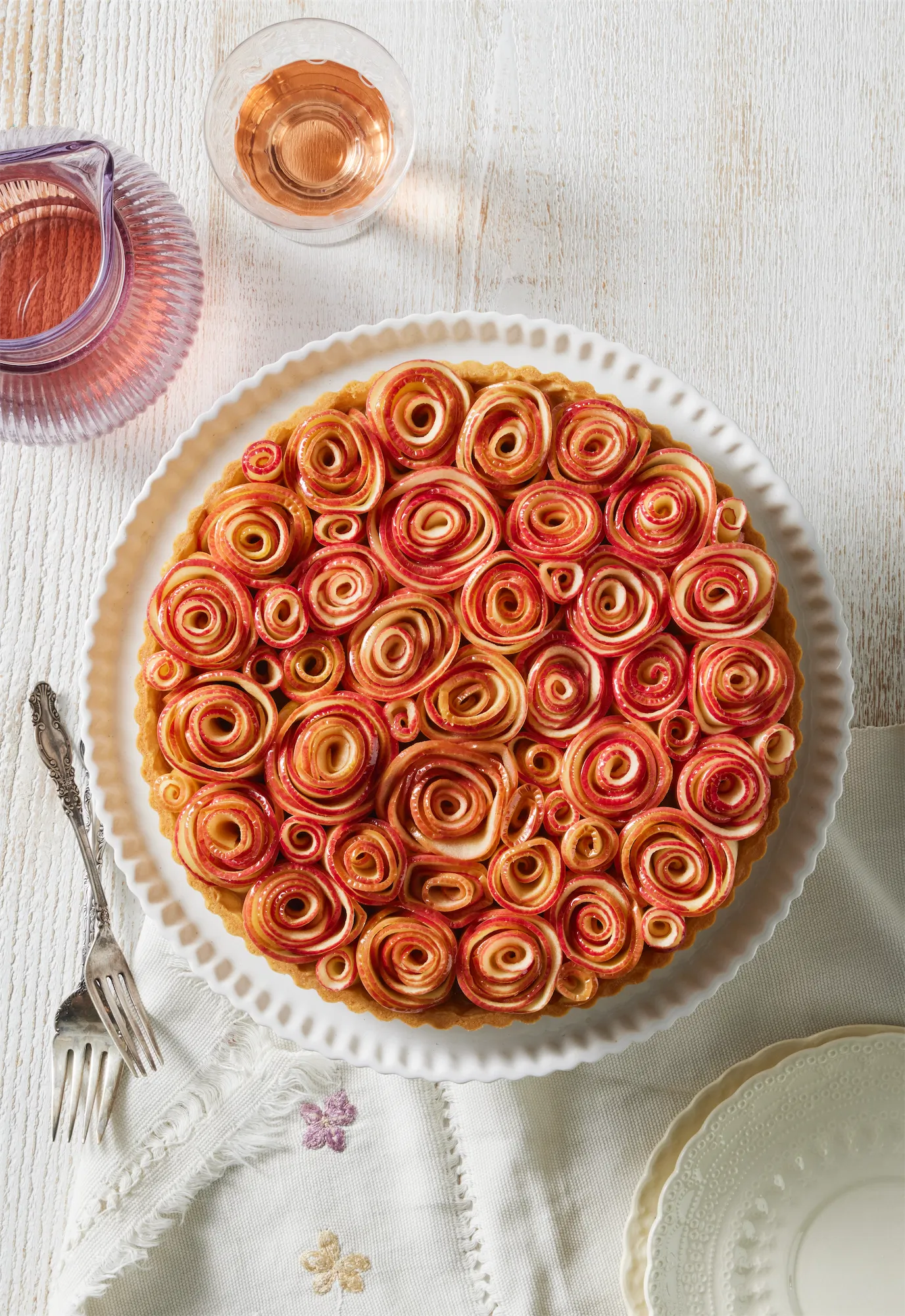 an overhead of apple tart made with roses of sliced apples a purple pitcher of rose wine and two forks to the left