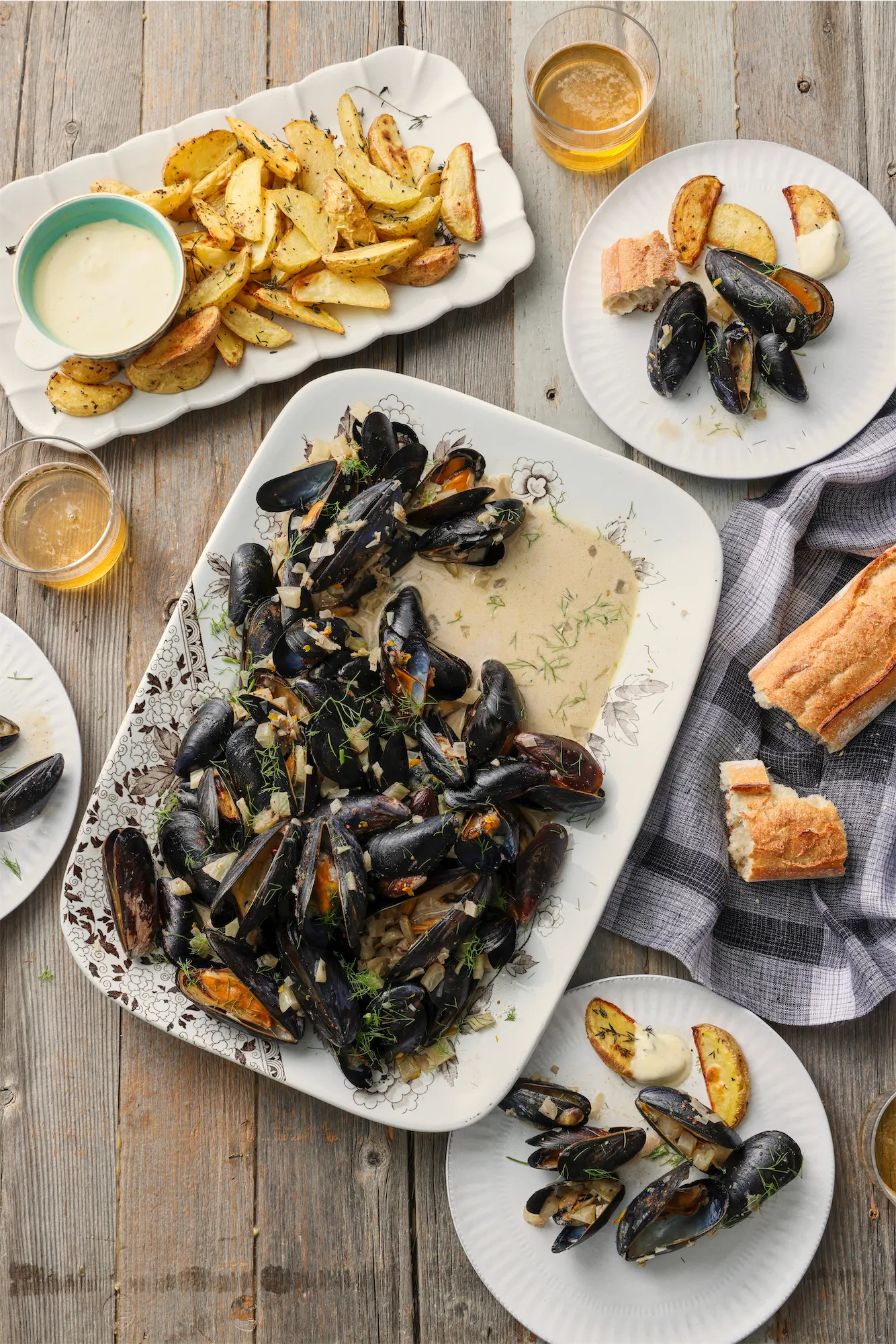 a large platter of steamed mussels in a creamy sauce with roasted potato wedges crusty bread and glasses of beer on a rustic table