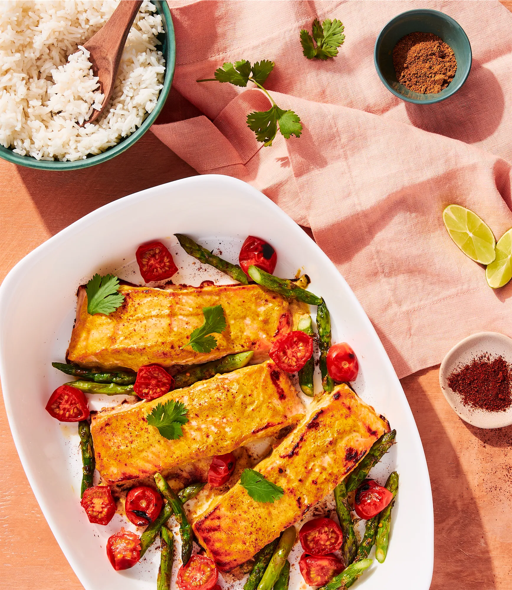 three salmon filets in a baking dish with cherry tomatoes asparagus and rice on a peach surface
