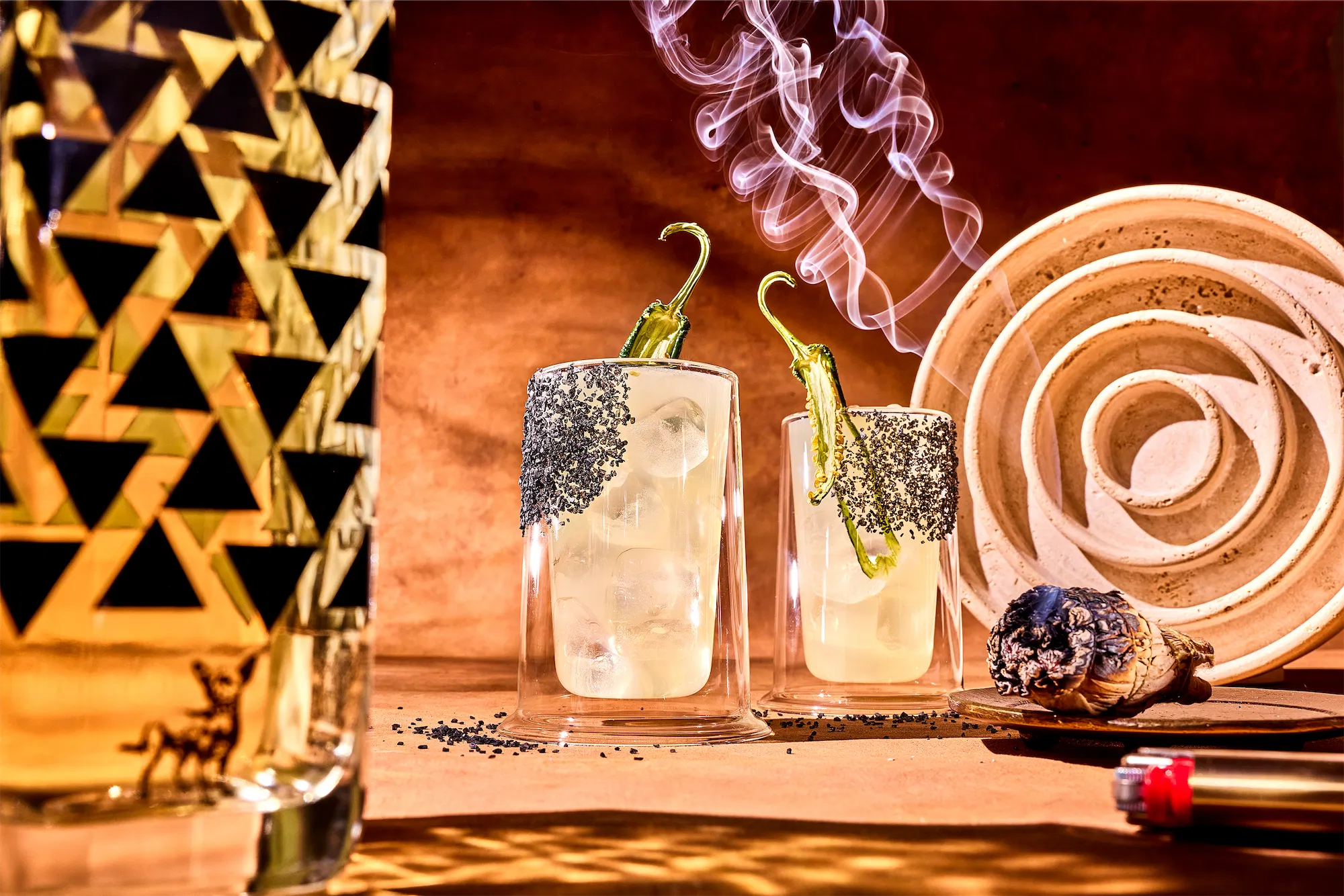 a surreal scene inspired by ai two cocktails rimmed with balck salt and jalapeno garnish a liquor bottle with black traingle pattern and a concentric circle plate a bic lighter in the foreground