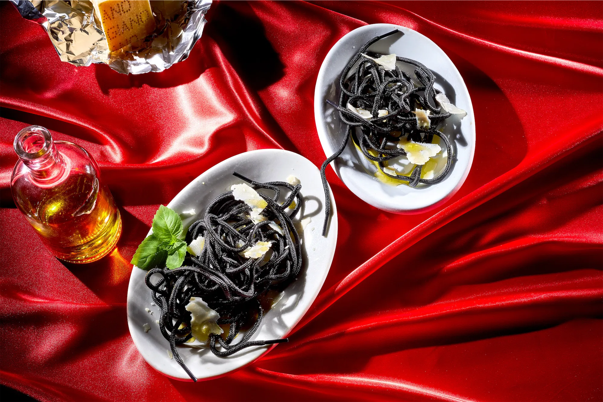 two white plates of shoelaces that mimic squid ink spaghetti garnished with fresh basil on a shiny red tablecloth with ruffled folds a bottle of olive oil and a parmesan reggiano wedge of cheese