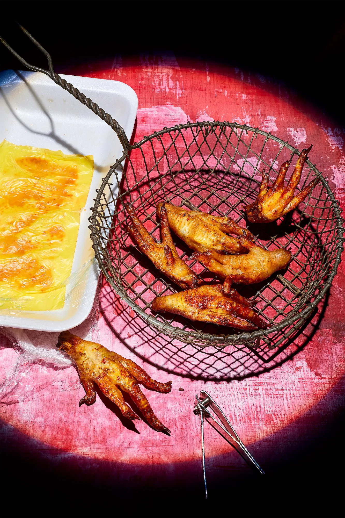 a spotlit scene of fried chicken feet ina metal basket on a pink surface with the polystyrene tray off to the left