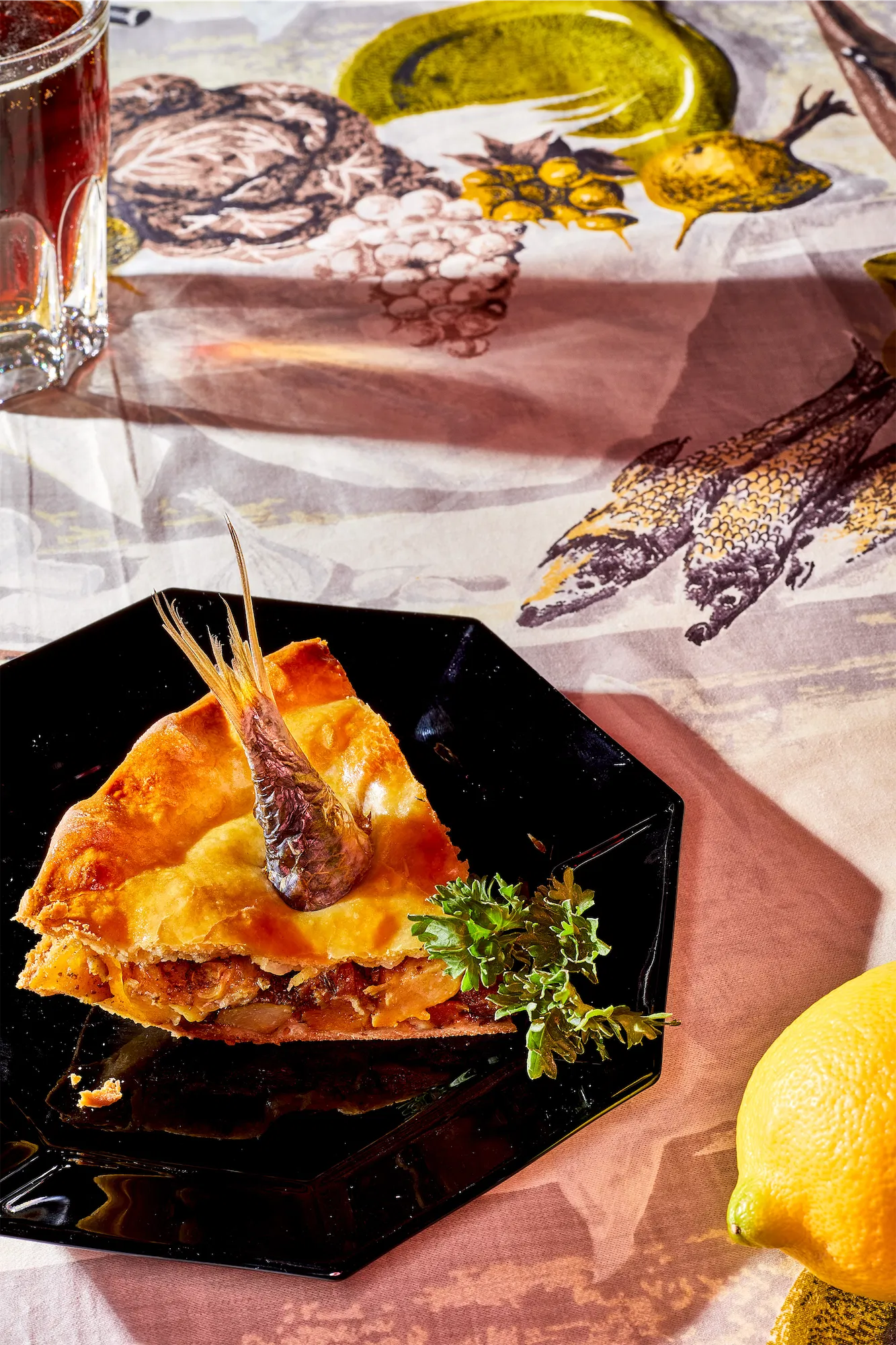 a slice of stargazey pie with a sardine tail on a black plate on a tablecoth with pictures of fish a lemon in the foreground