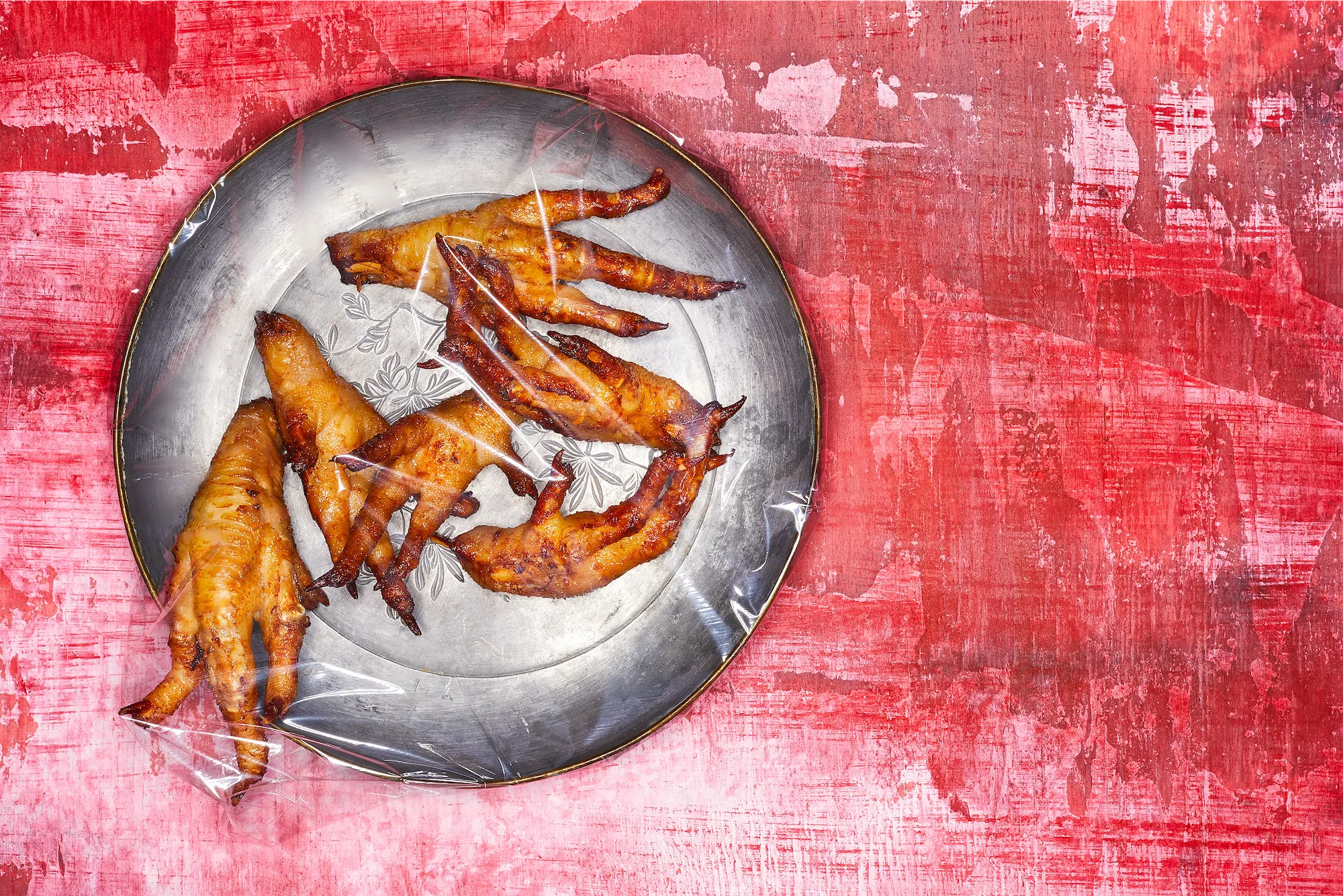 a pewter plate full of fried chicken feet covered in plastic wrap on a disterssed red surface