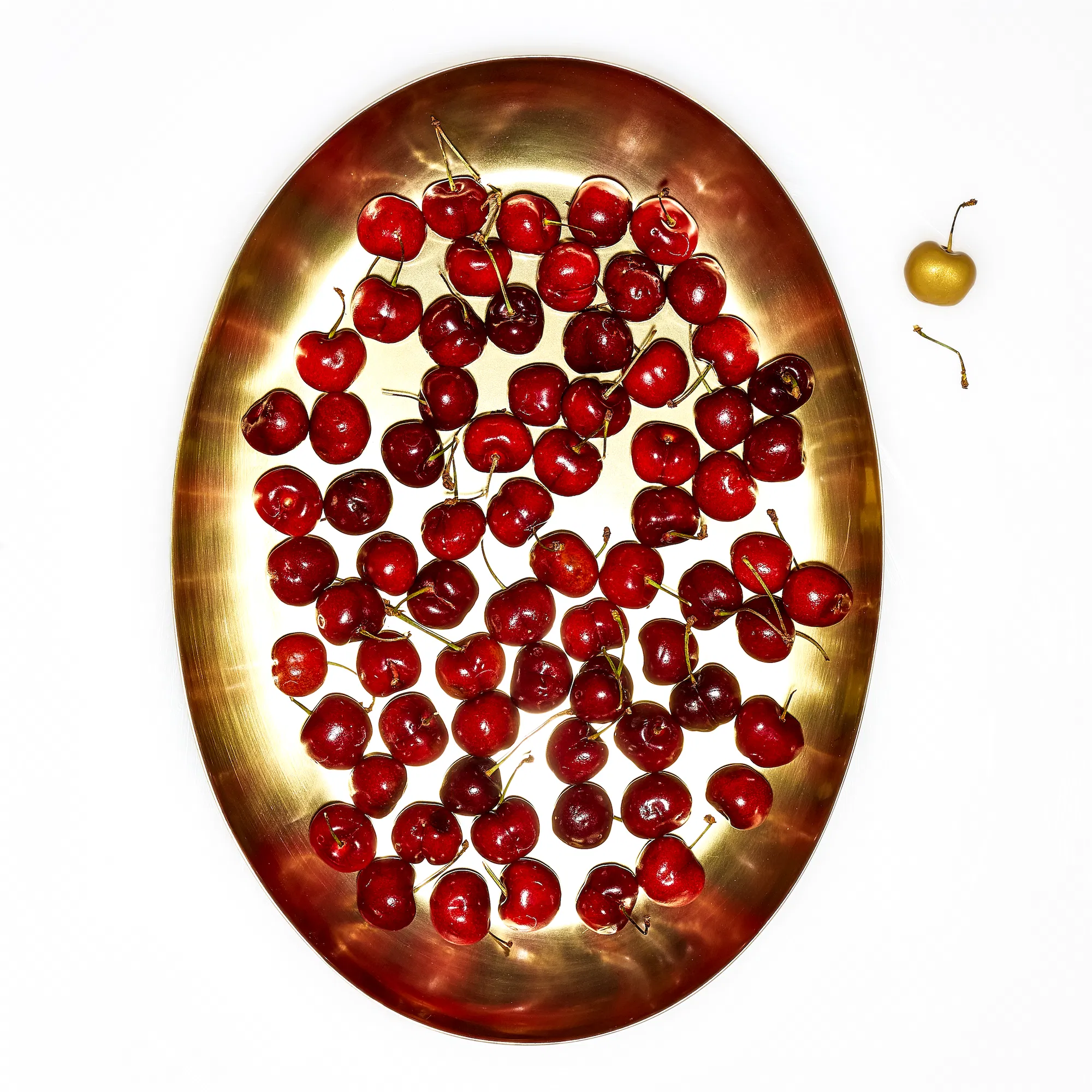 a gold tray full of red cherries with one gold cherry off to the side on a white background