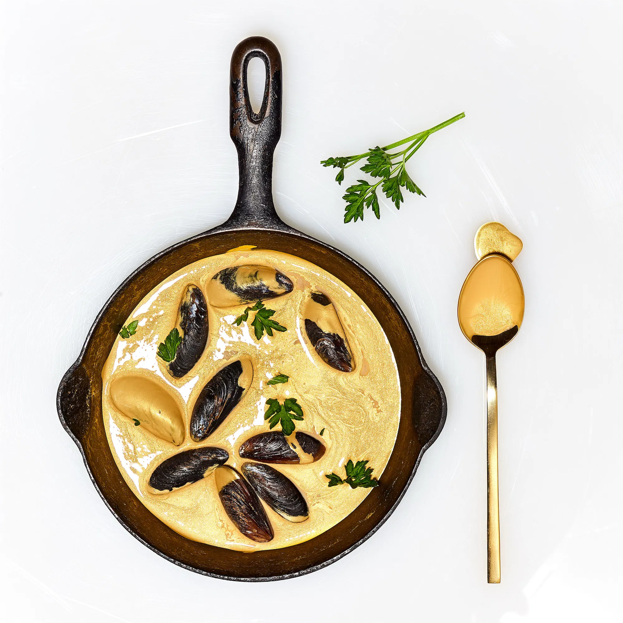 a cast iron skillet with mussels swimming in metallic gold paint ralph lauren brand parsley garnish and a gold spoon to the right on a white background