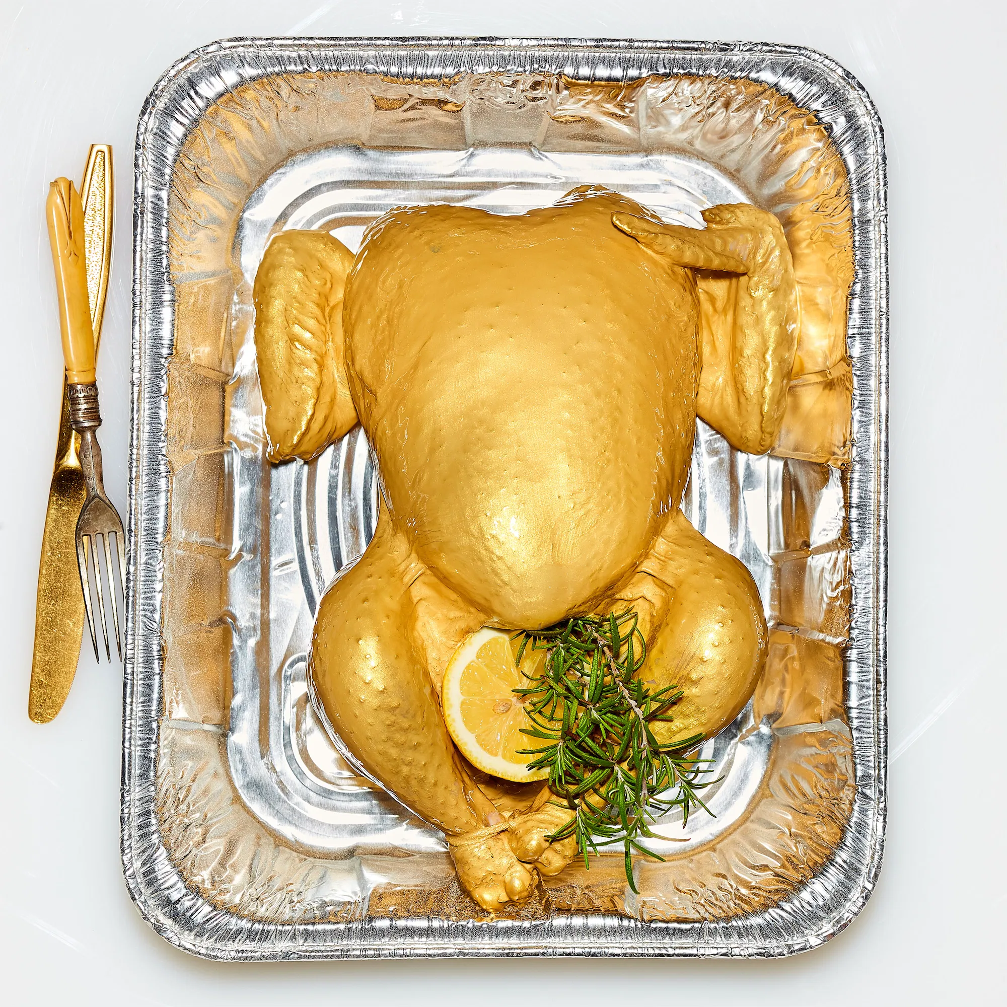 a gold chicken in a metallic silver roasting tray with rosemary lemon garnish and upside down vinatge knife and fork to the left