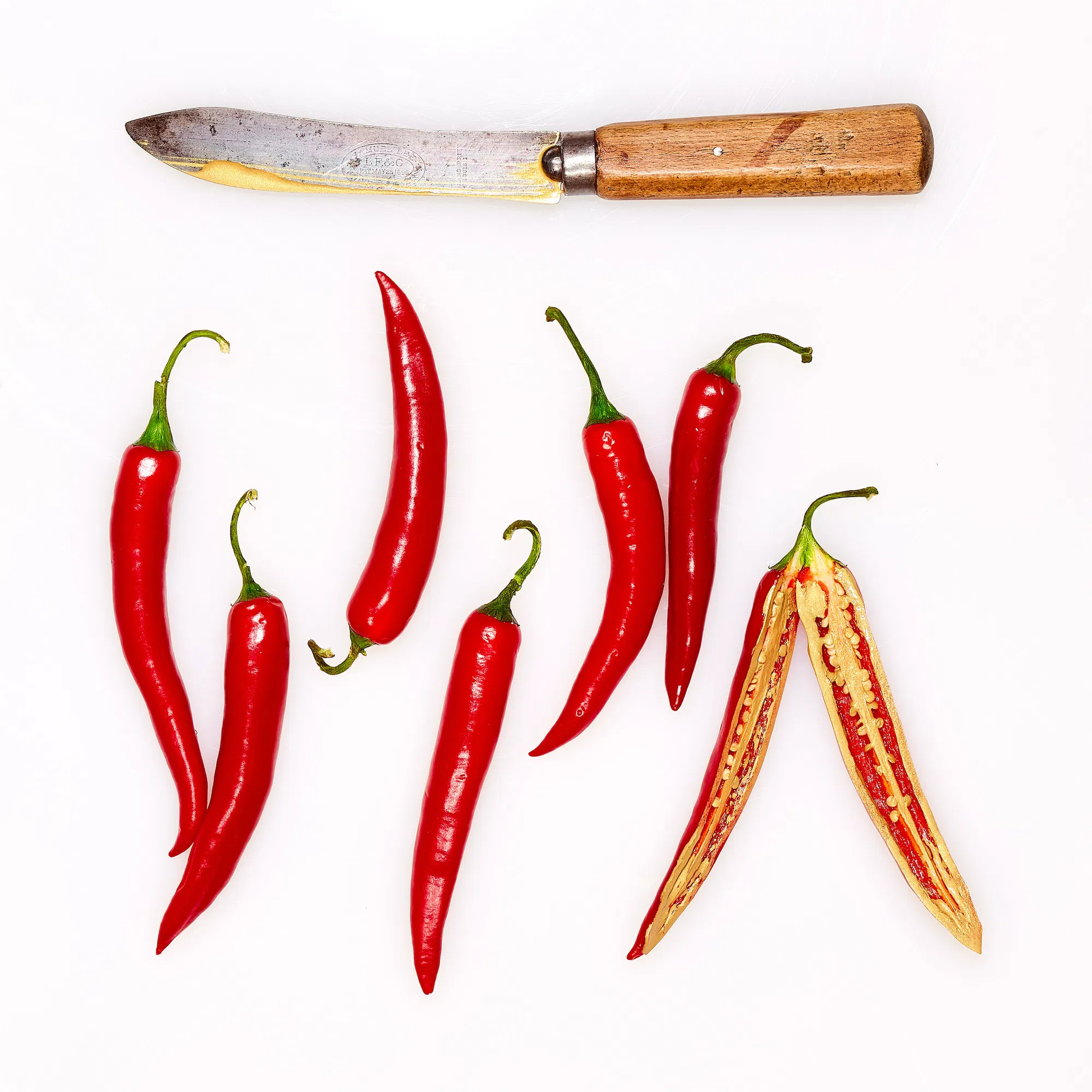 6 red chilis and one gold chili cut in half with a gold dipped vintage knife above on a white background