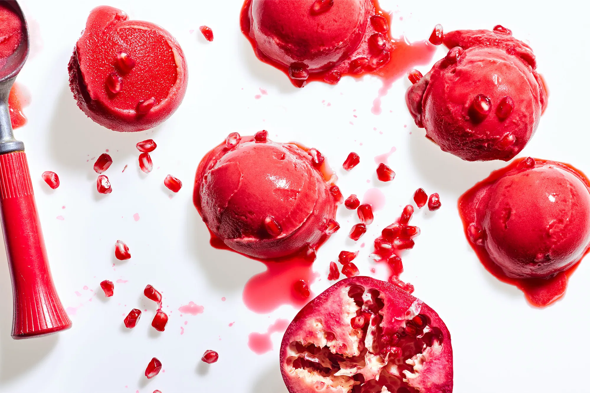 5 scoops of red sorbet ice cream and a red handled scoop and half a pomegranate with the arils scattered on a white background