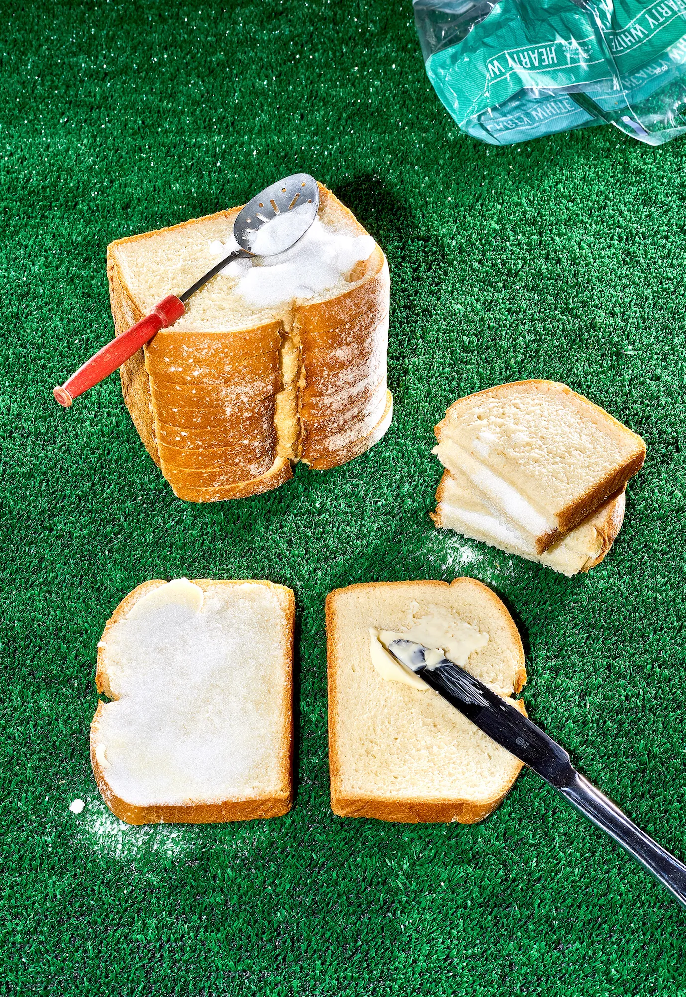 a stack of white bread with a sugar sandwich being made on a fake grass surface with a red handled spoon and metal knife