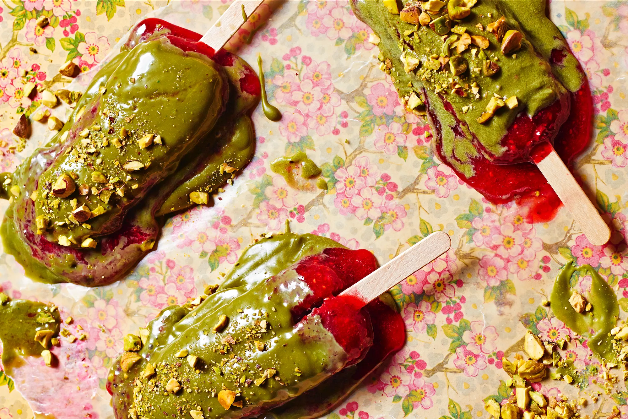 3 ice creams on sticks melting onto a floral surface with pistachio green coating and red revealing