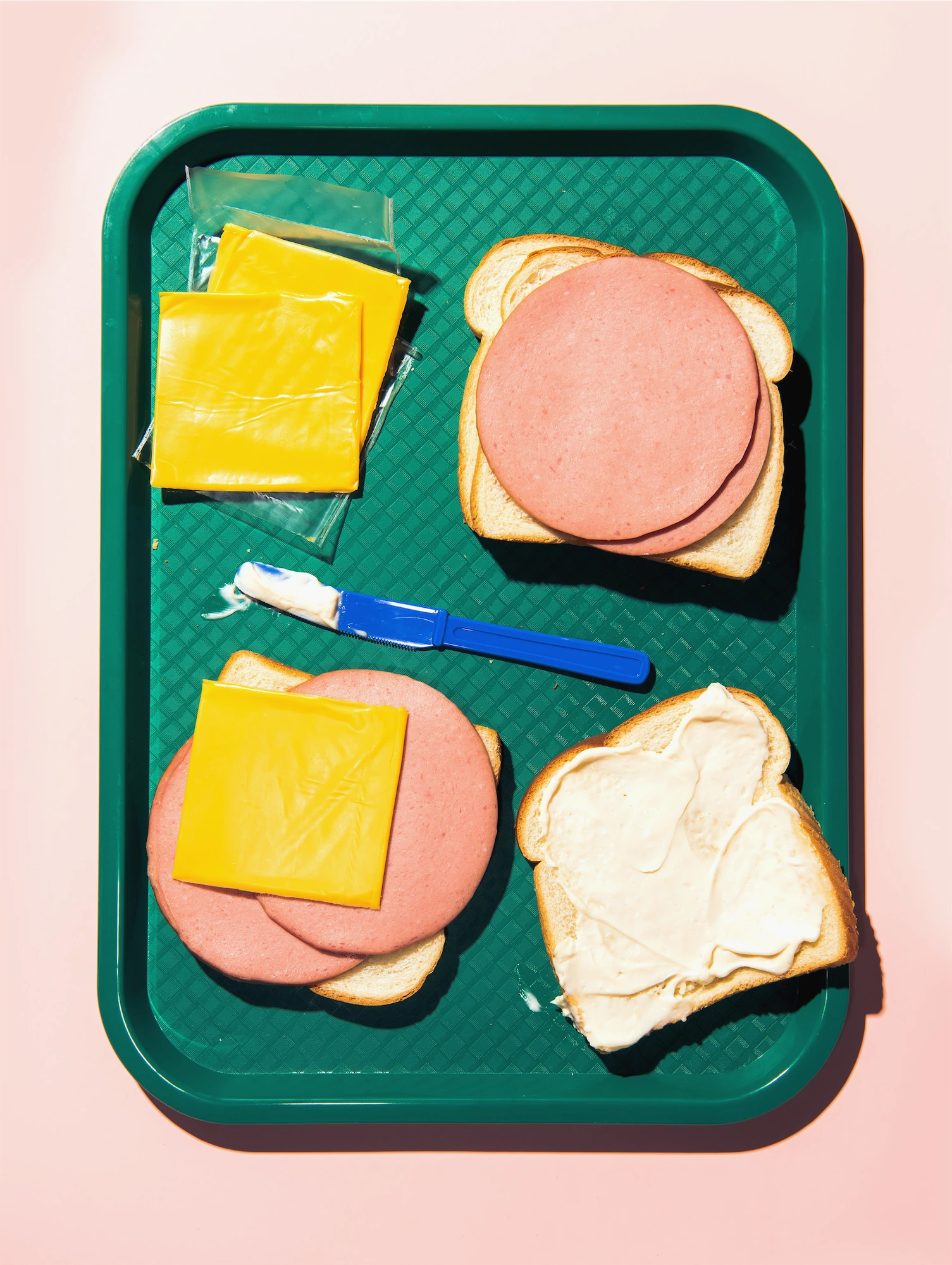 a green canteen tray with the makings of a baloney sandwich in progress ameriacn cheese slices and mayonnaise with a blue plastic knife