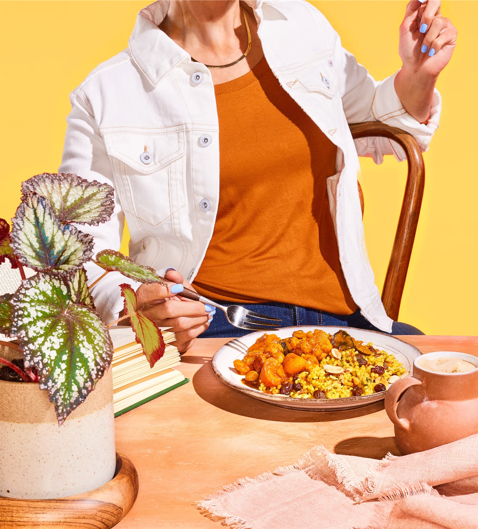 world fare brand scene of a model against a yellow background seated at a table with a plate of food and a houseplant in the foreground