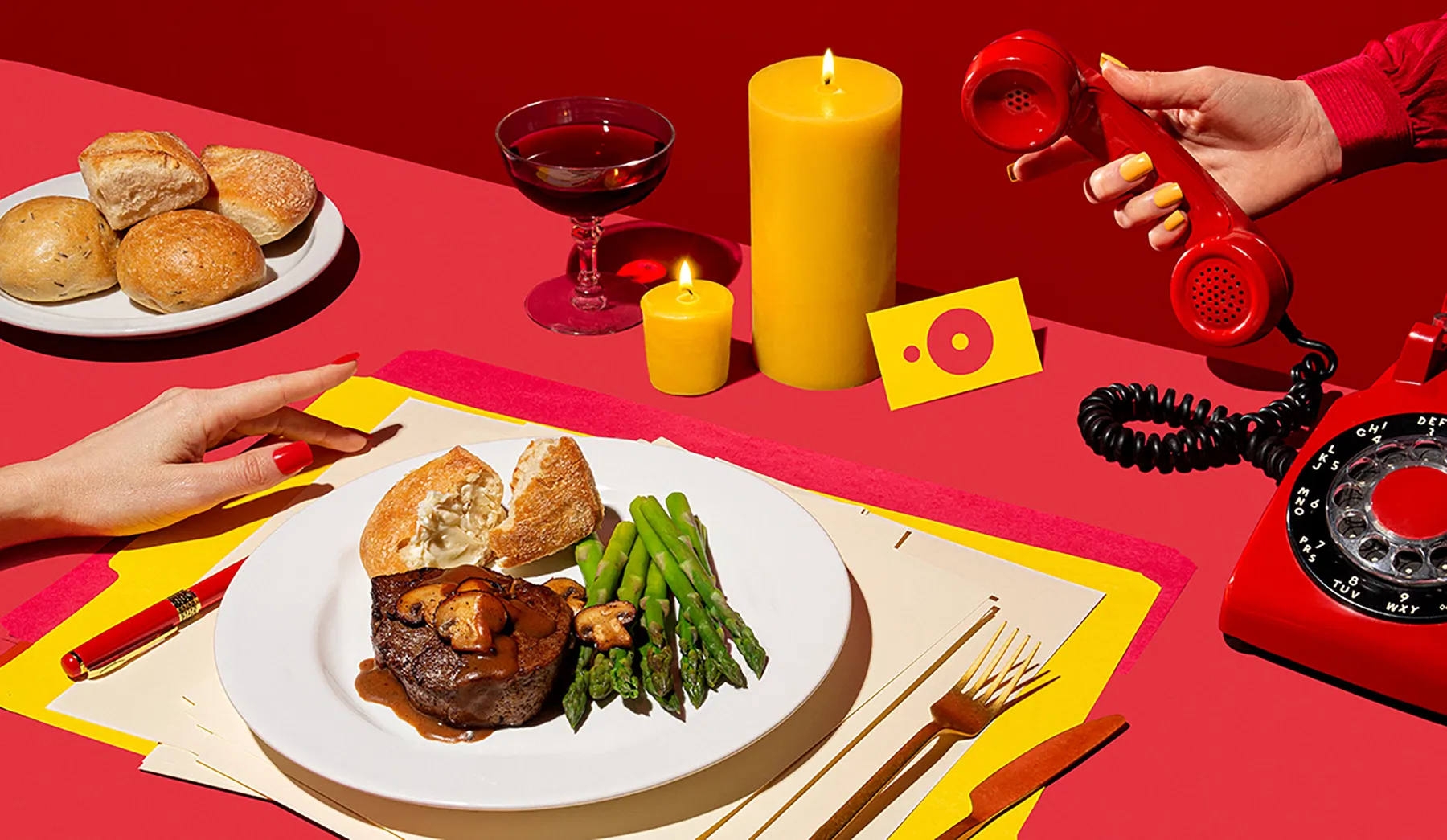 a red and yellow scene of a hand beside a plate of filet mignon with asparagus and bread rolls another hand holding a red telephone off the hook with a yellow candle burning