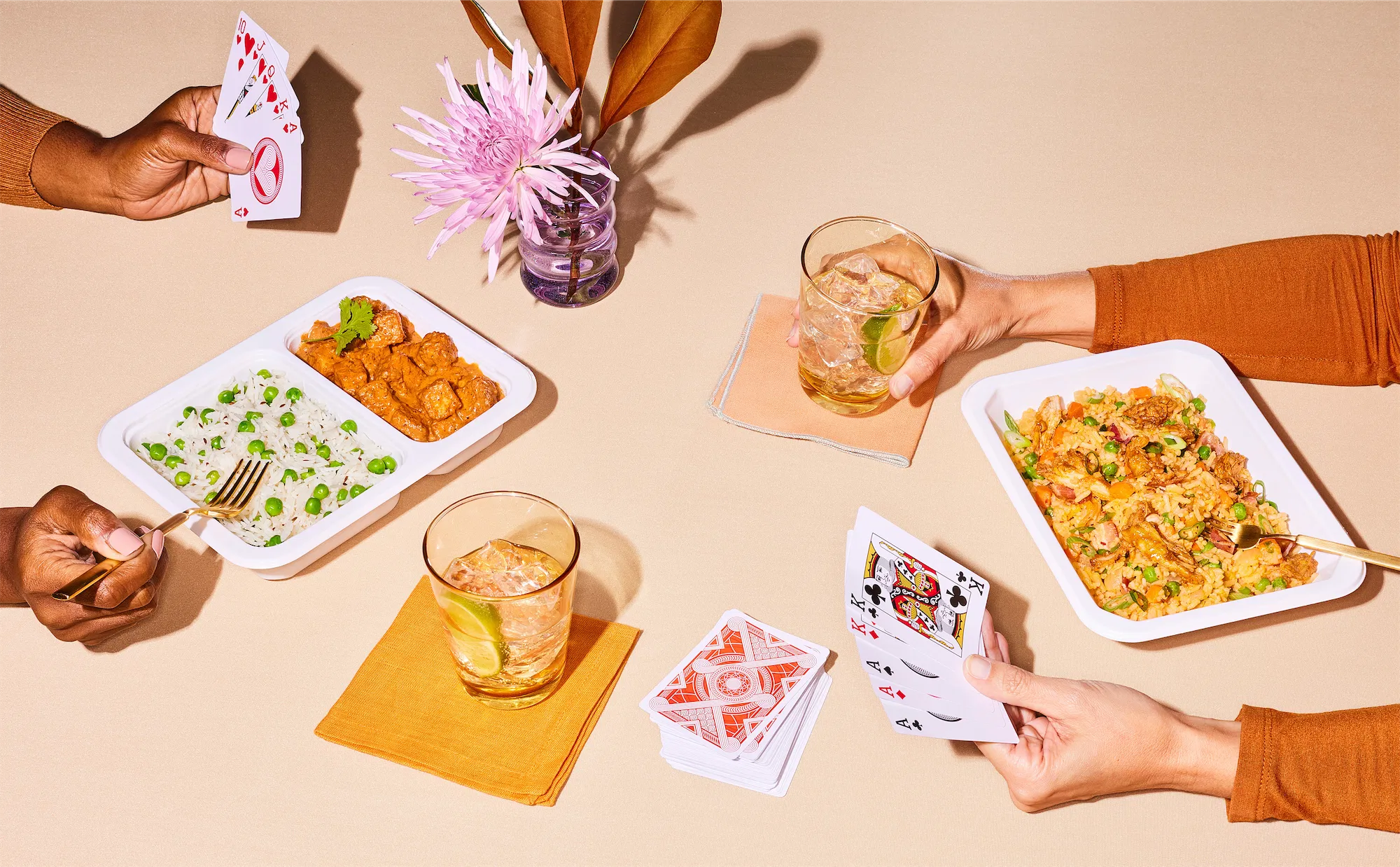 two pair of hands sit playing cards at a table with ready meals and a large crysanthemum in the center two iced drinks on the table