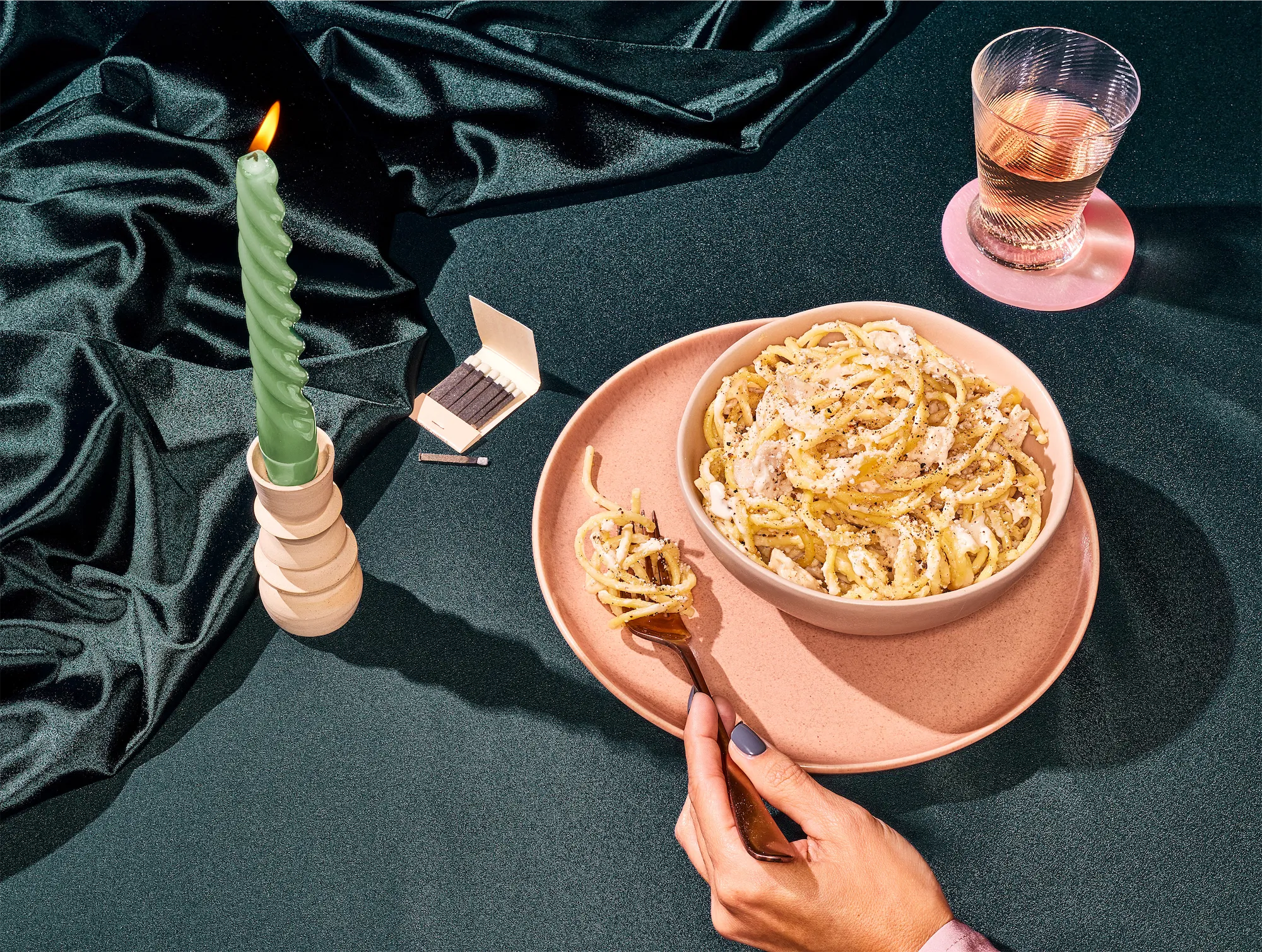 on a black ruffled fabric background sits a bowl of pasta on a similar colored plate with a hand holding a fork while a green ribbed candle flickers