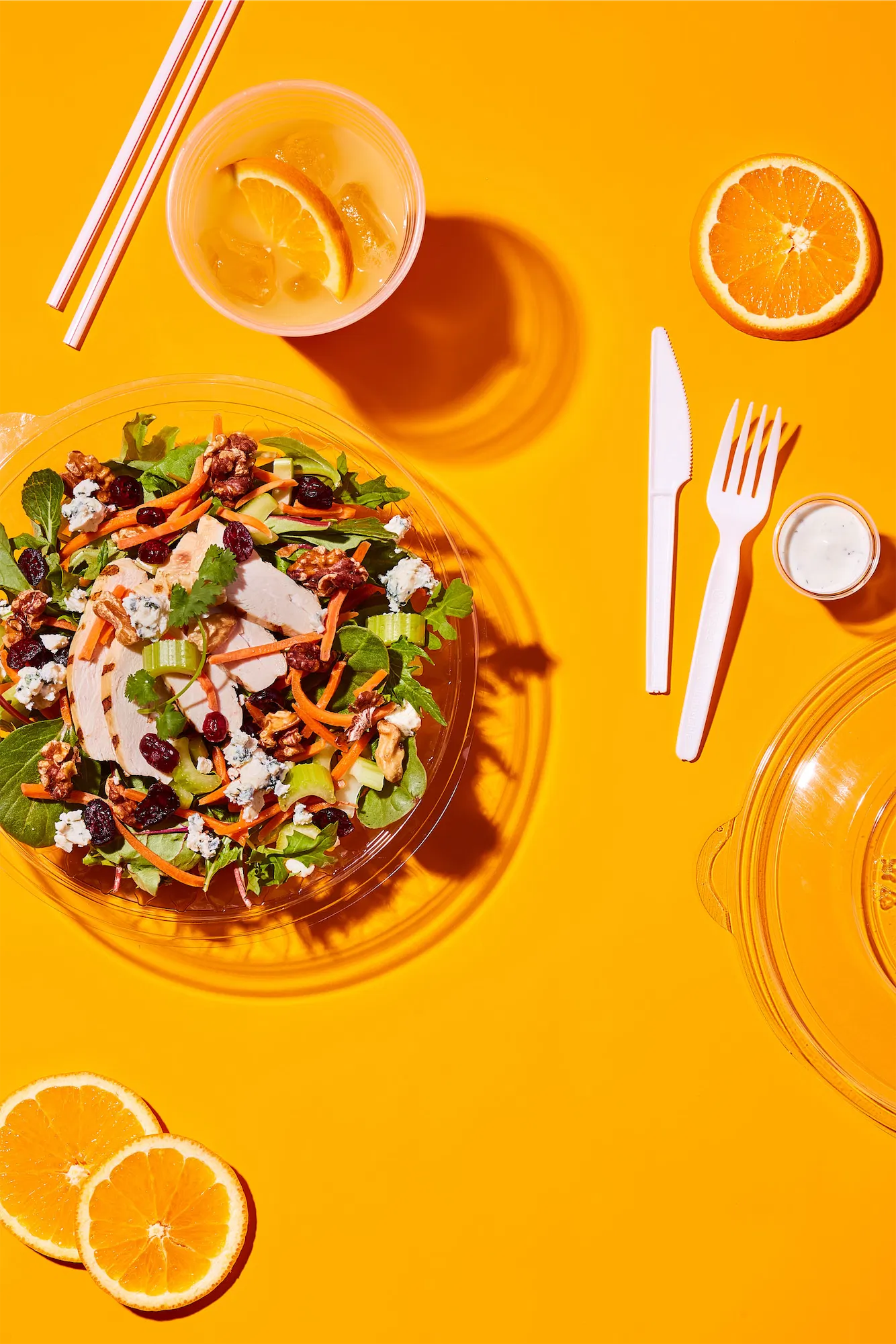 a chicken salad in a transparent plastic take-out container on an orange background with white plastic utensils and an orange juice with two straws