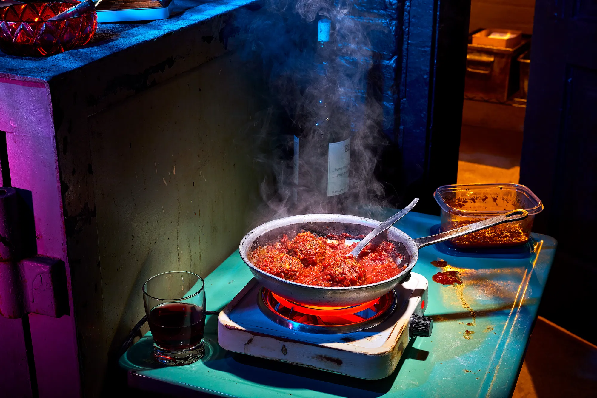 a nightime scene with a pan of simmering meatballs in red sauce on an electric plate with steam rising and a glass of red wine