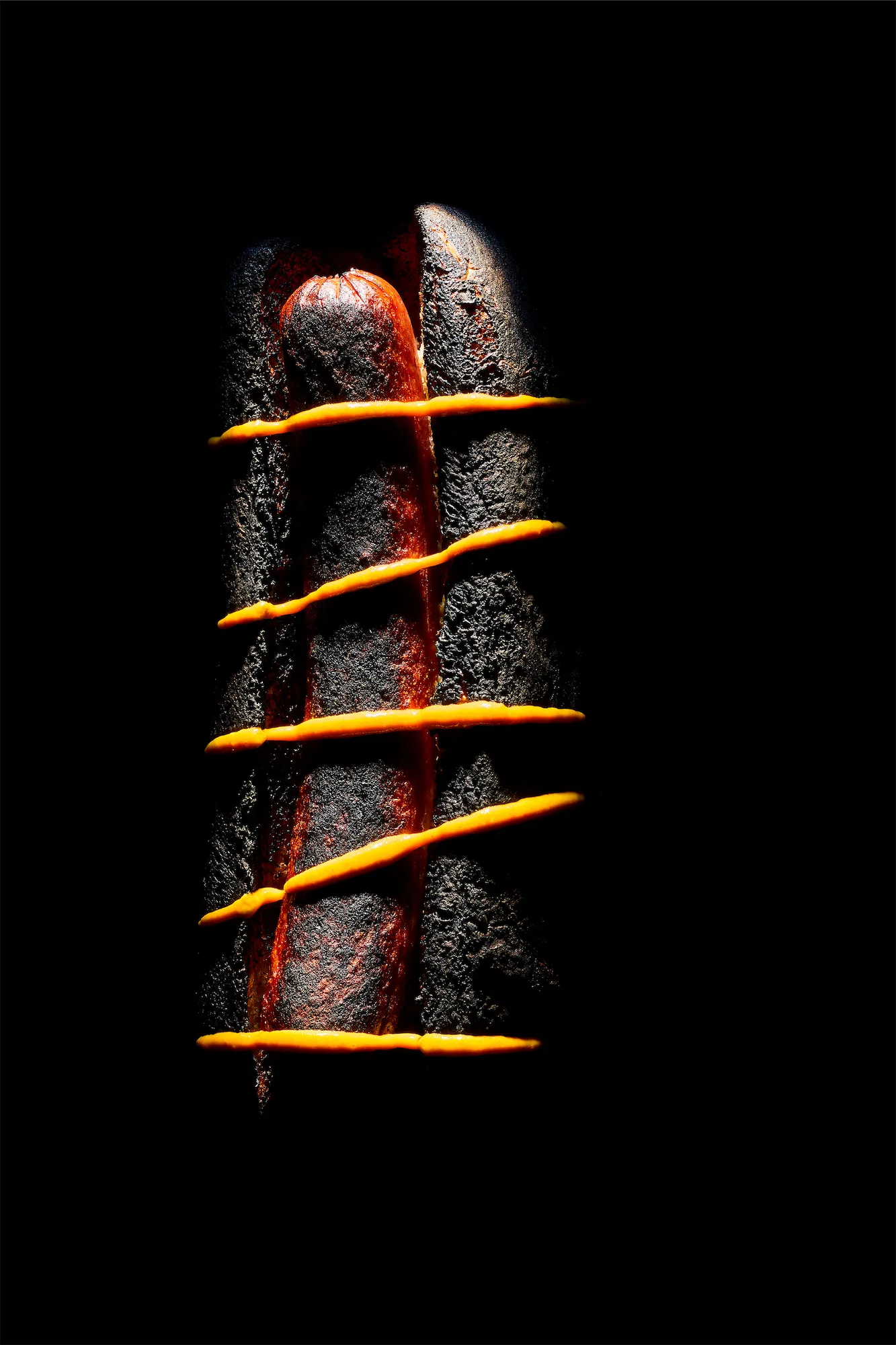 a blackened hotdog with lines of american mustard across it on a black bckground