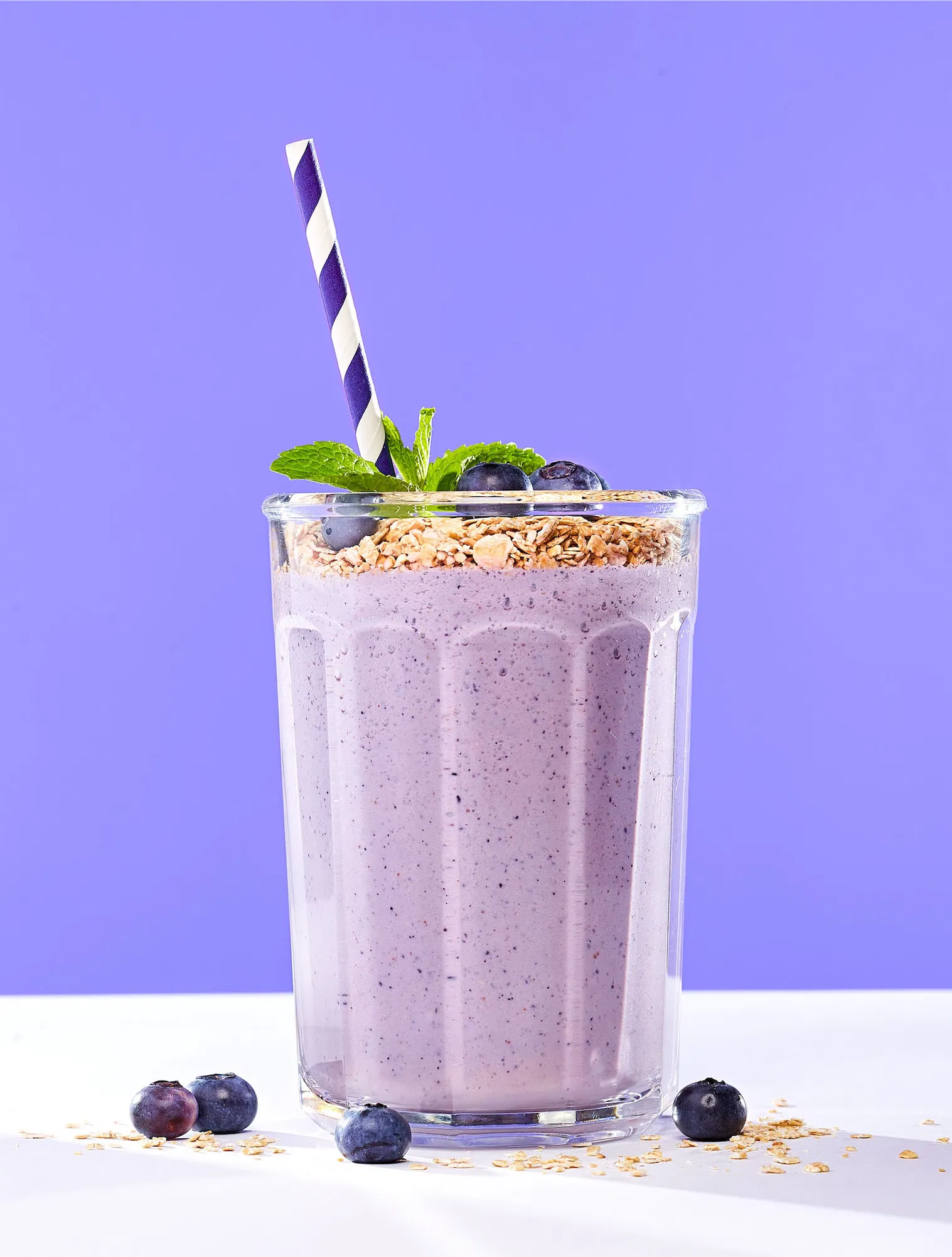 a blueberry smoothie in a tall ribbed glass with a blue striped straw against a purple background