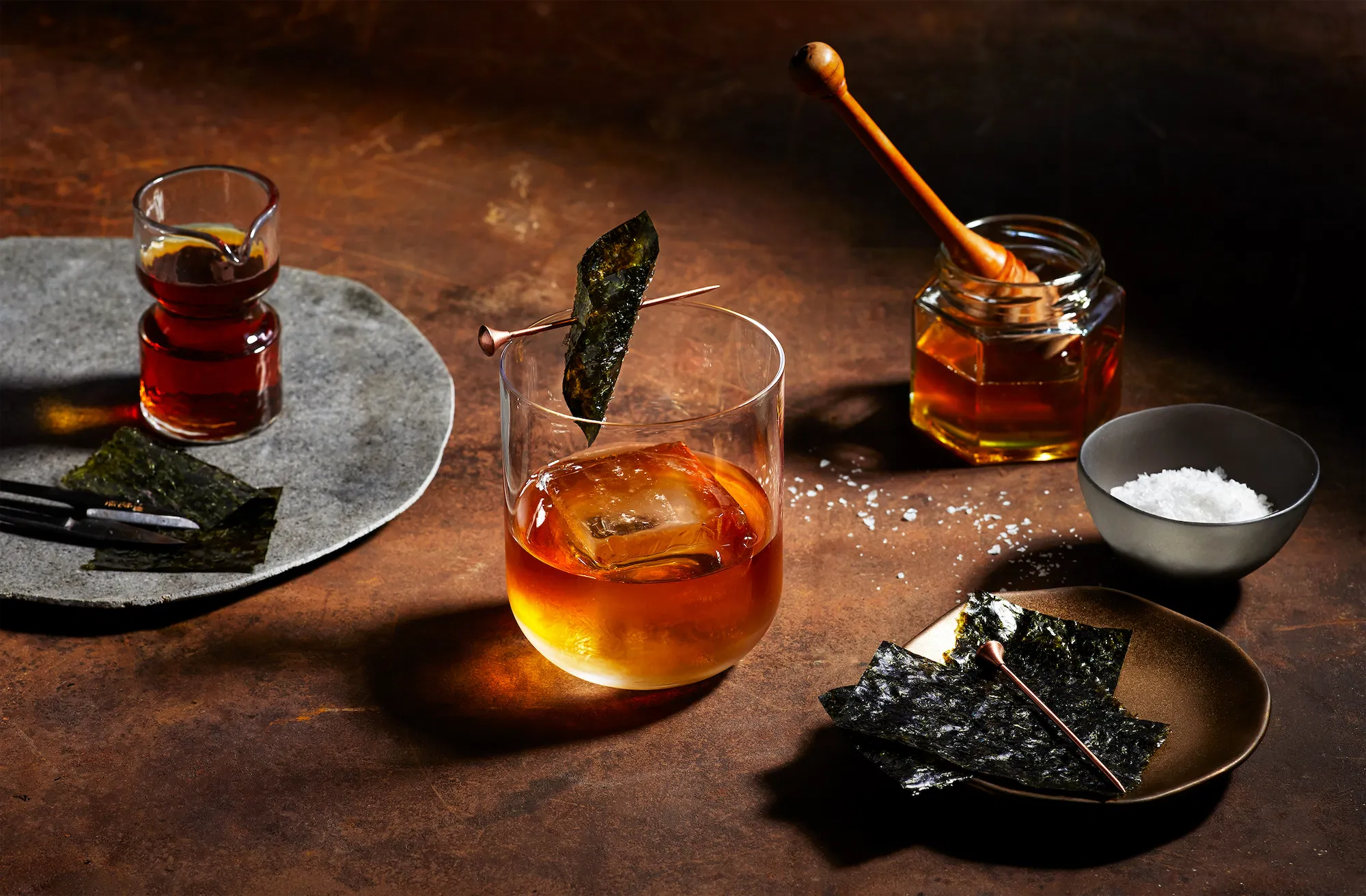 bourbon old fashioned in a rocks glass on a brown surface with honey and seaweed garnish