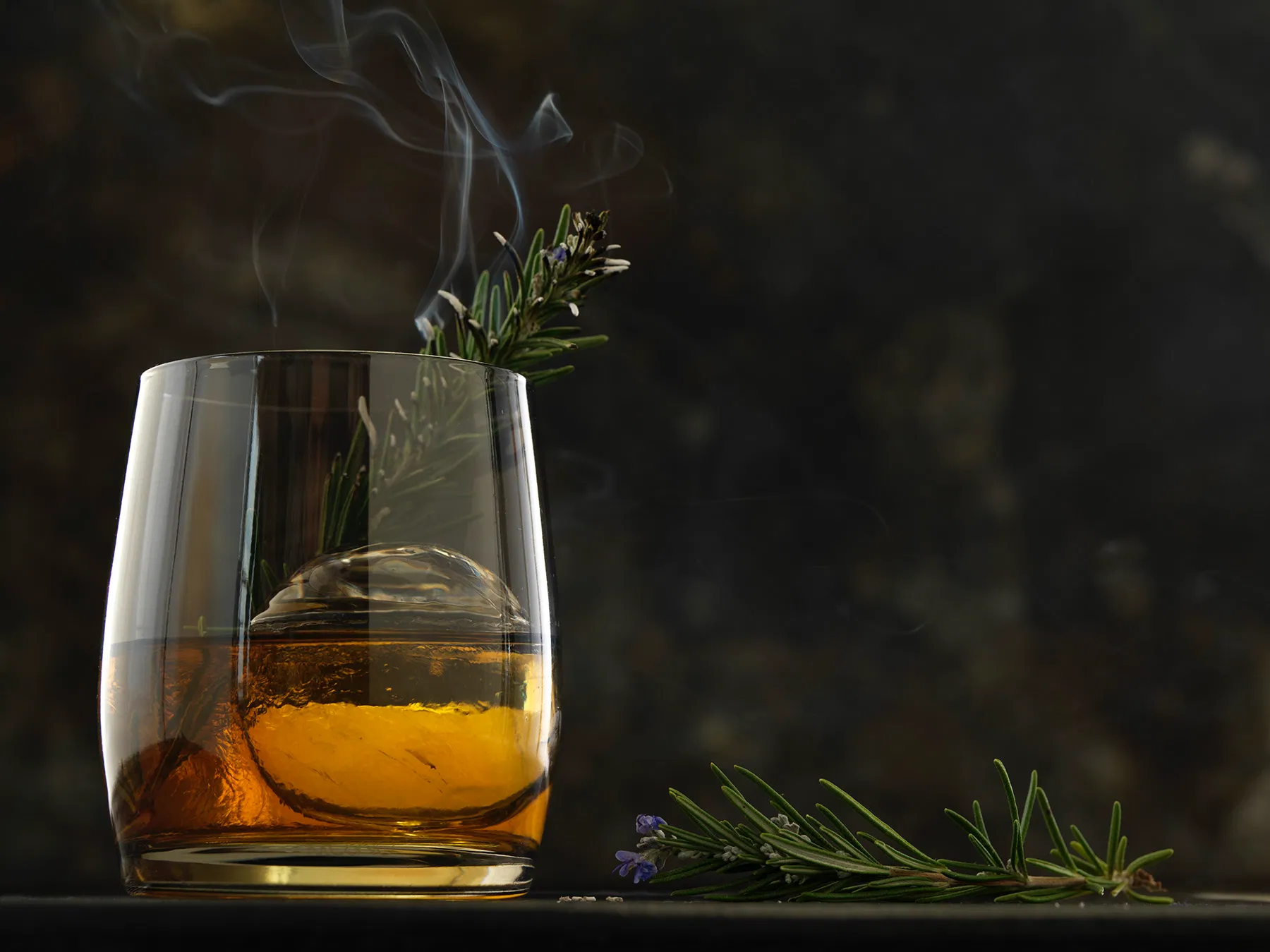 Bourbon old fashione in a rocks glass with large round ice cube and scorched rosemary garnish against moody dark background