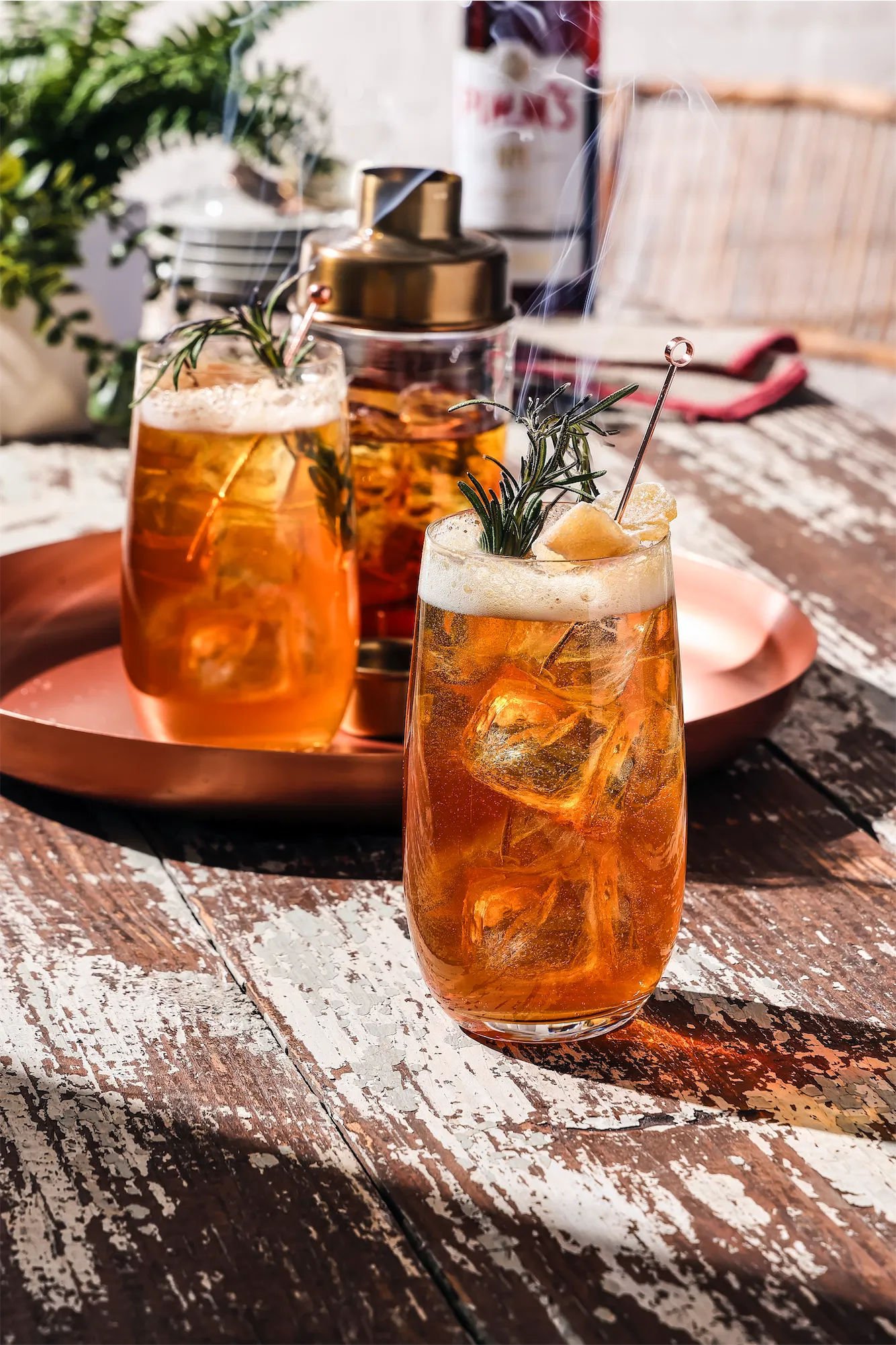 Pimm's cocktail in a tall glass on a sunny table with rustic vibe and smokey scorched rosemary as a garnish with lemon peel