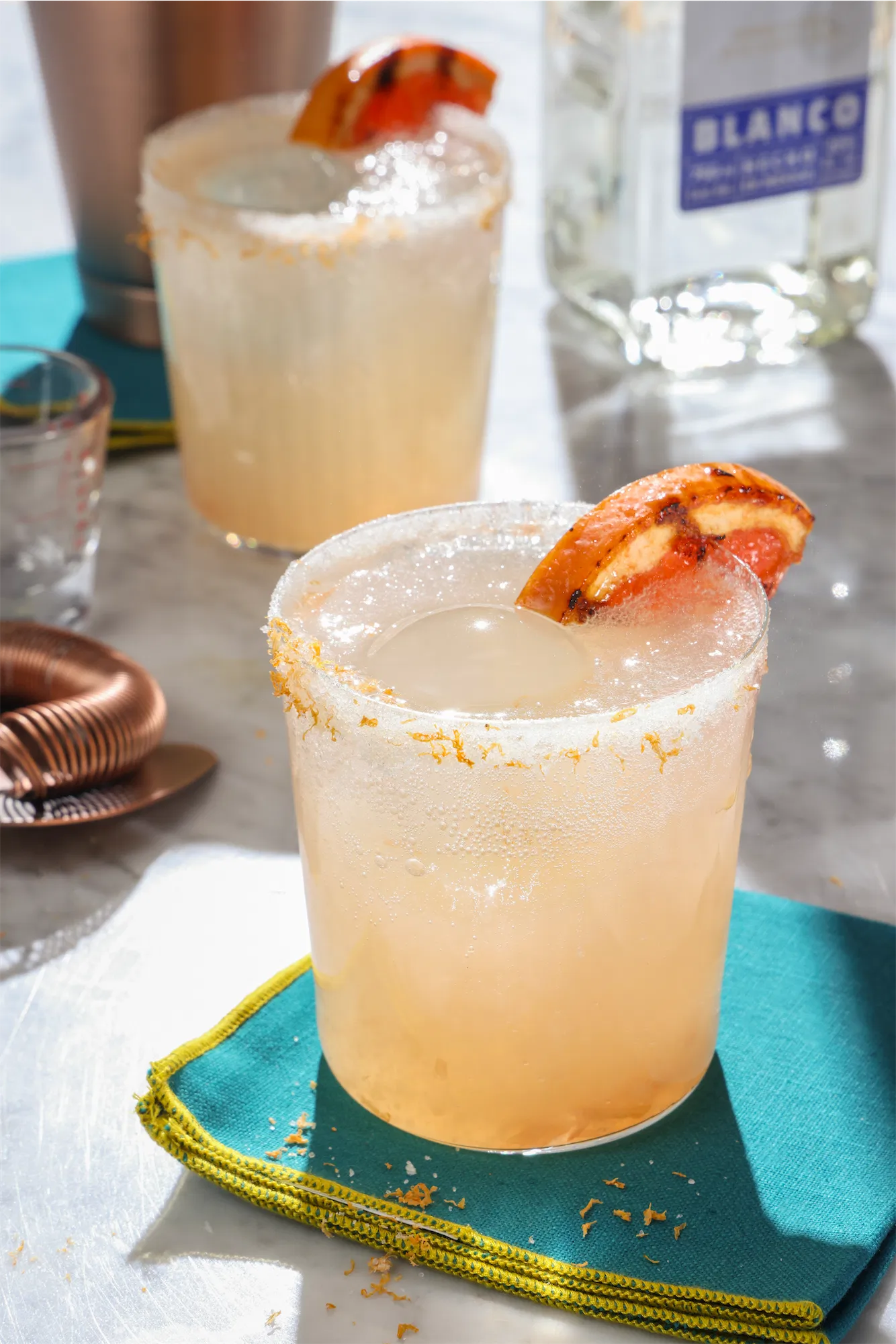 sunny scene showing sugar rimmed cocktail glass filled with a grapefruit paloma made with tequila and garnished with a grilled orange slice
