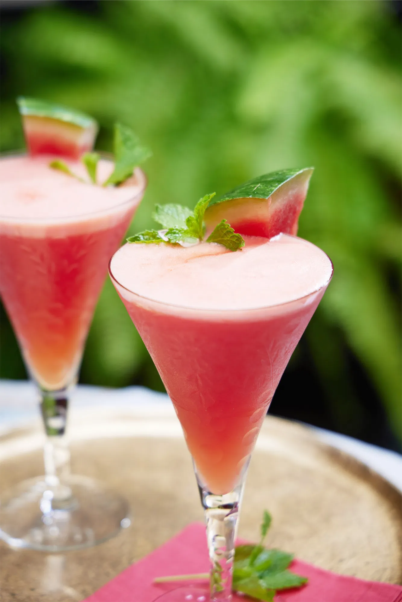a watermelon daiquiri in a martini glass with mint garnish on a gold tray against a green fern backdrop