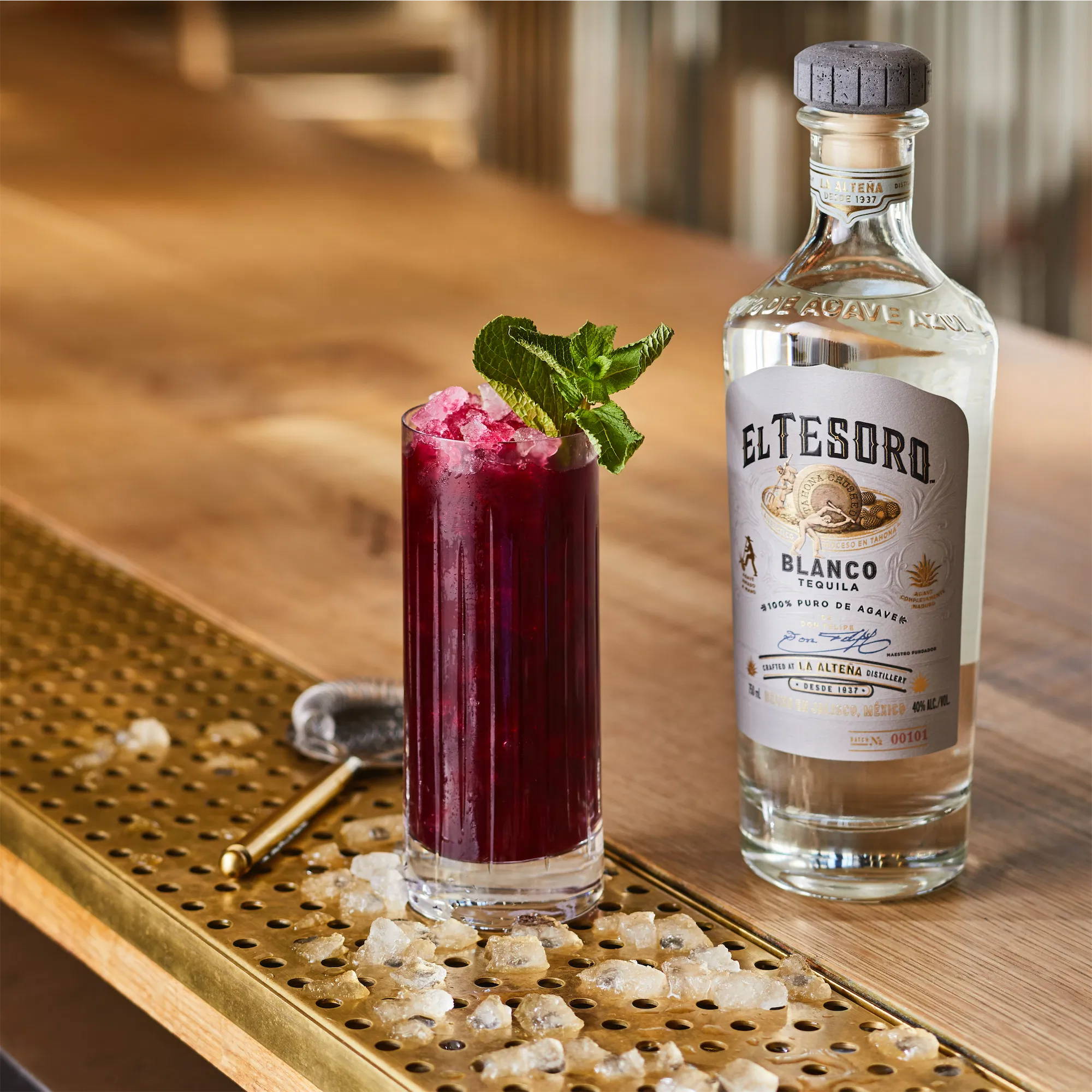 white label brand El tesoro blanco tequila bottle on a bar top with a burgundy colored cocktail in a collins galss surrounded by ice garnished with mint