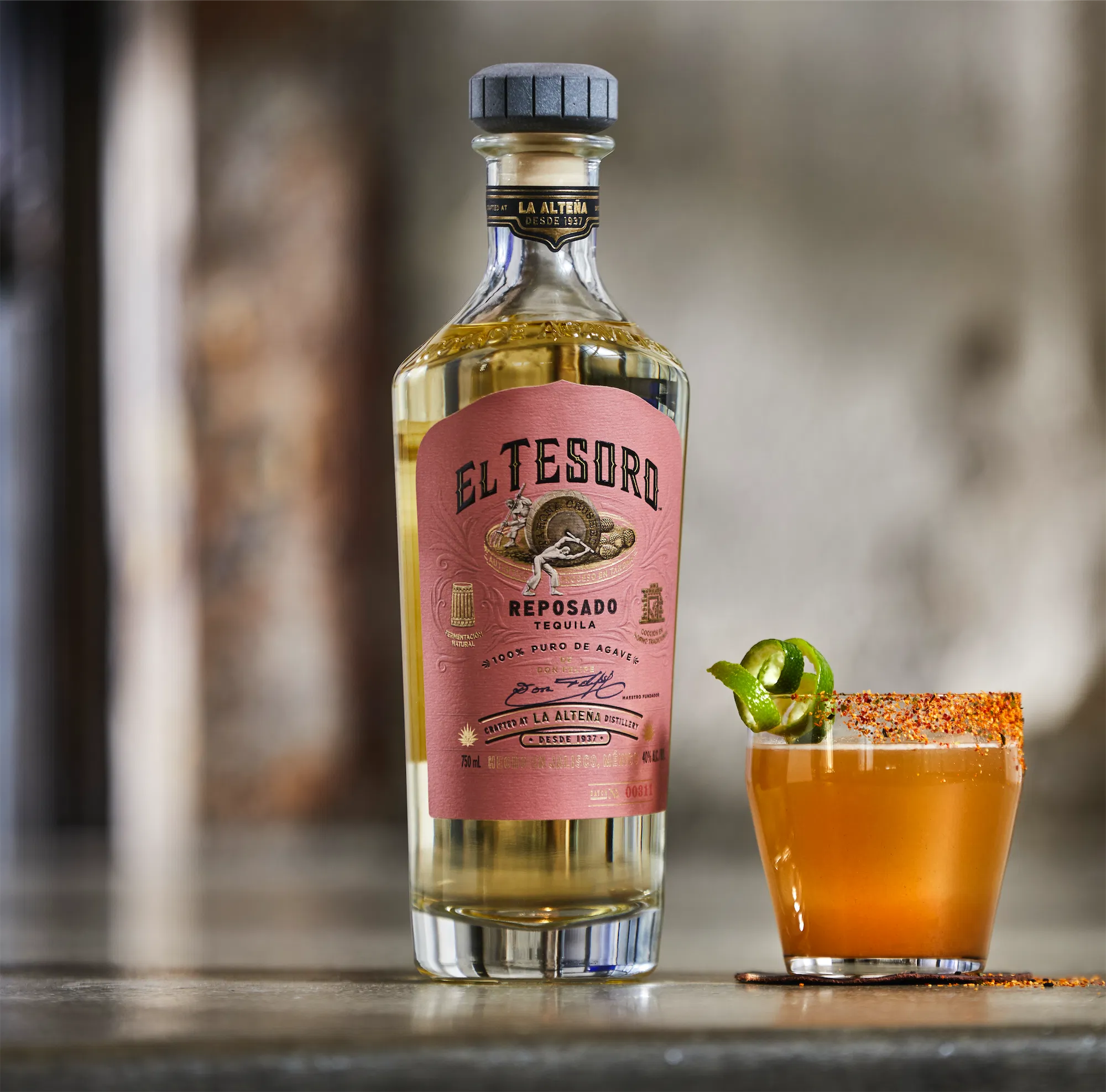 White label brand e tesoro reposado tequila bottle next to rocks glass filled with a spicy cocktail rimmed with chili powder and garnished with jalapeno