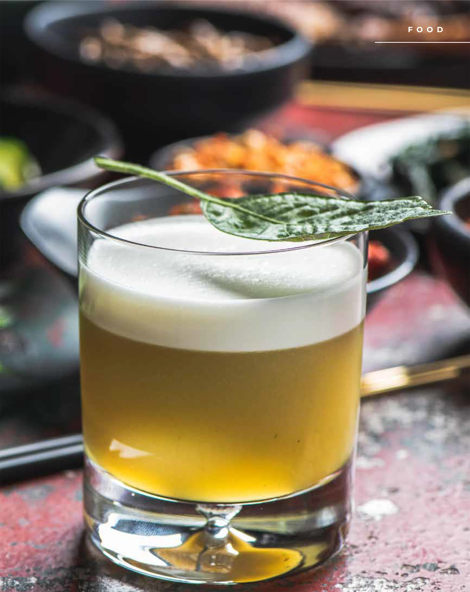 yellow cocktail with a foam head garnished with perilla leaf