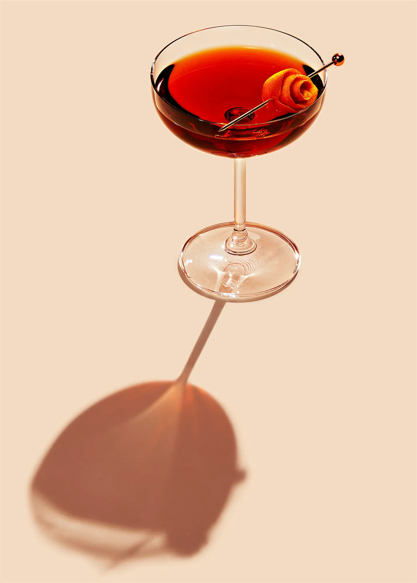 Jim Beam cocktail in a coupe garnished with orange twist on metal pick and a long shadow on plain background