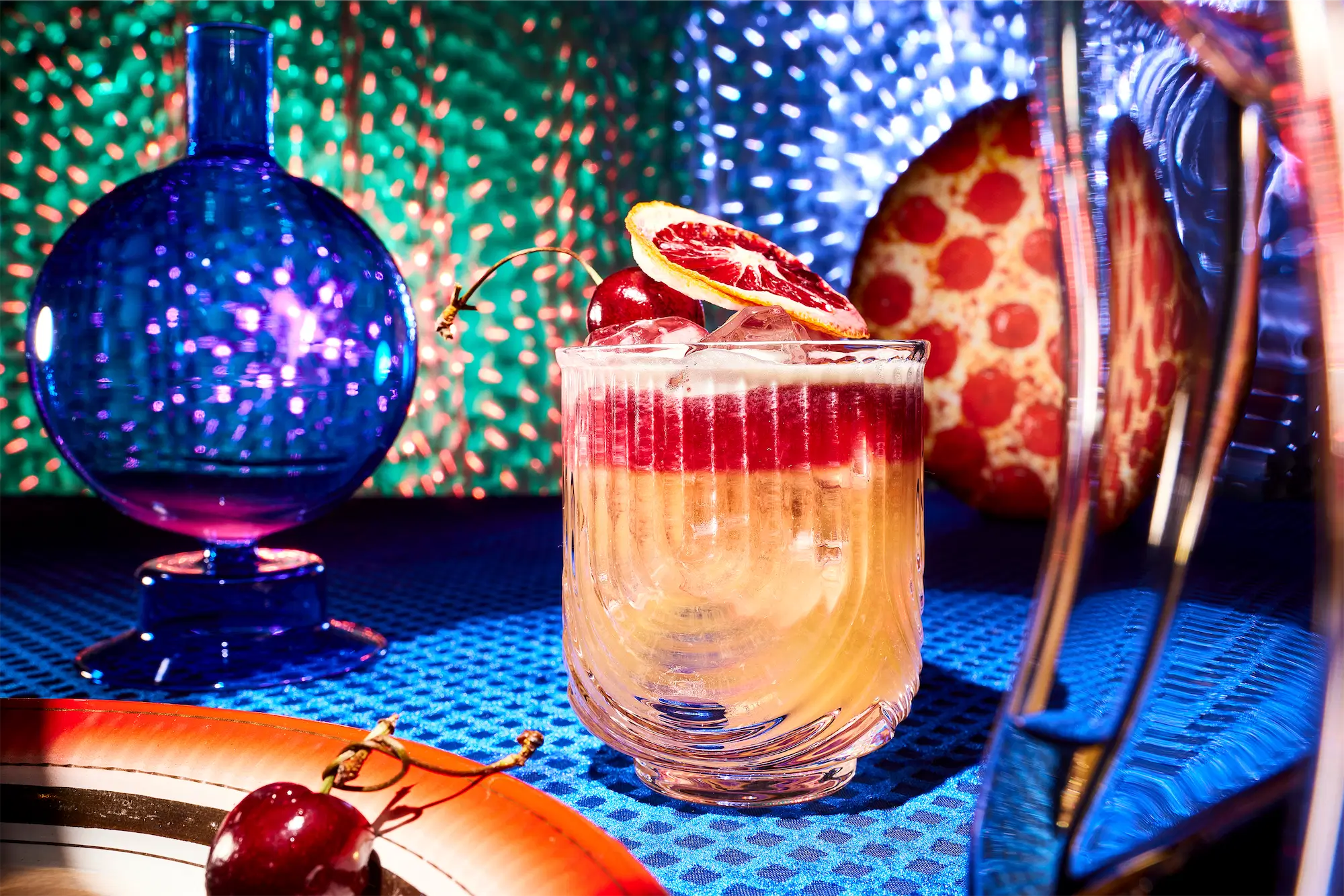 a surreal scene insopired by ai of a layered cocktail in a ribbed glass garnished with grapefruit on a blue patterened tablecloth a pizza in the background and a disco lit wall a cherry in the foreground