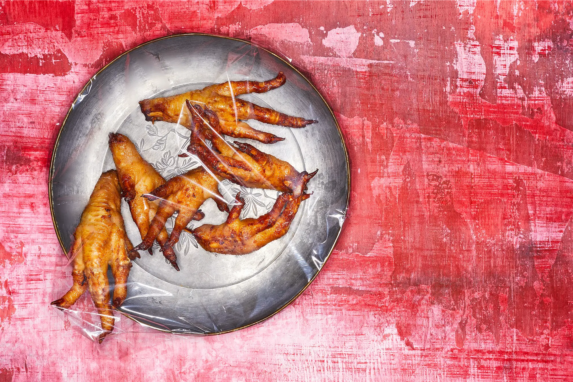 a pewter plate full of fried chicken feet covered in plastic wrap on a disterssed red surface