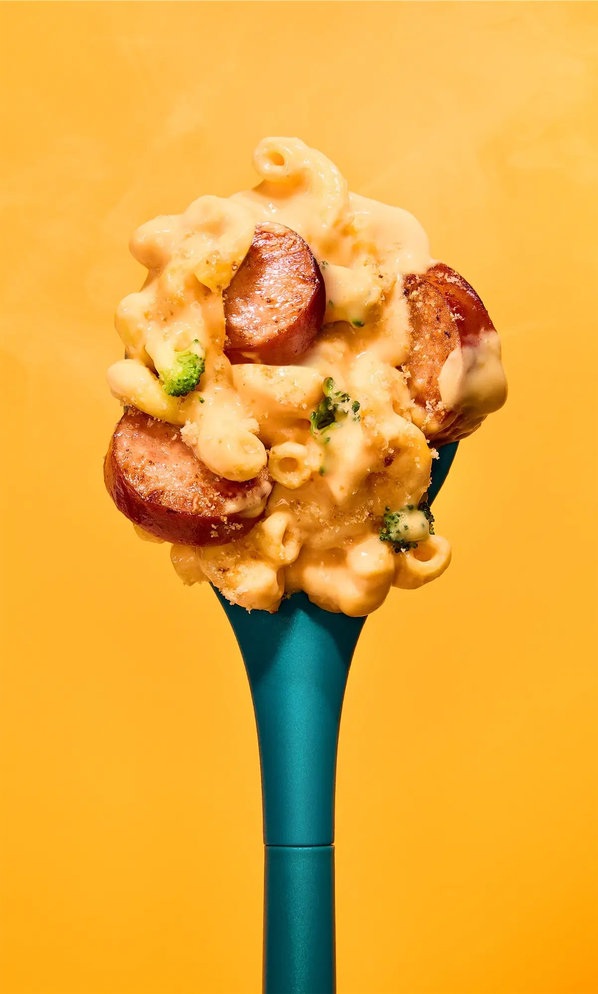 Johnsonville close-up of creamy mac and cheese with smoky sausage slices and broccoli on a teal spoon against a yellow background